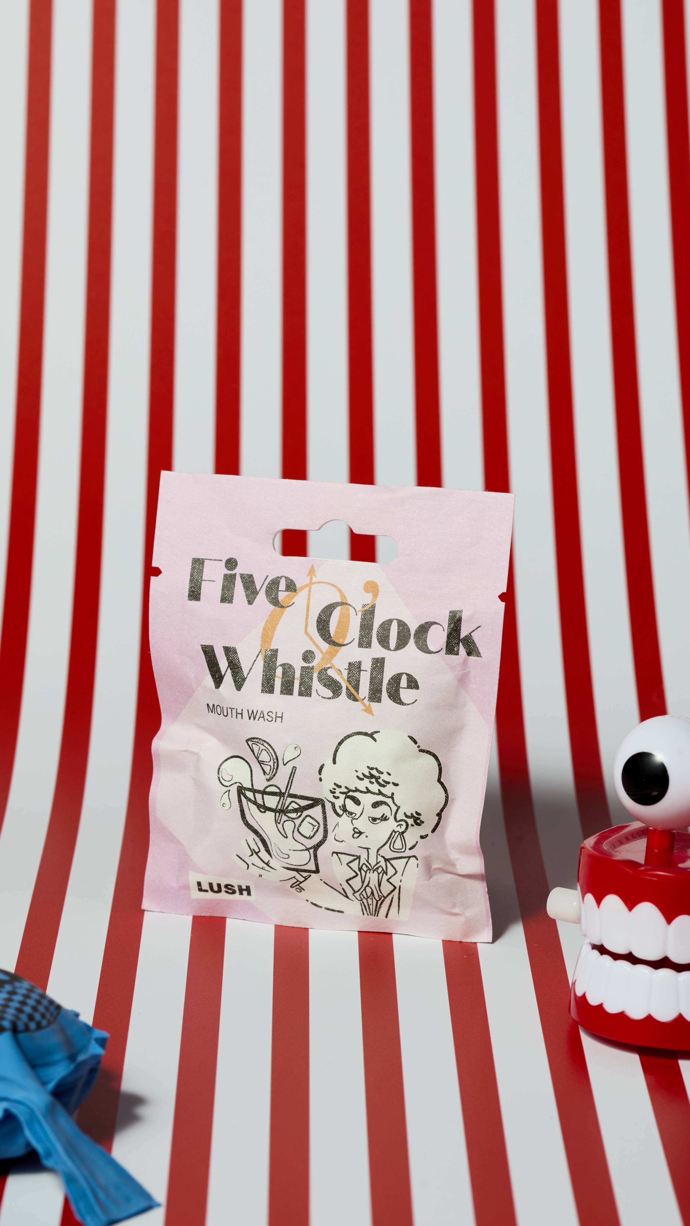 The image shows the Five O'Clock Whistle Mout Wash product sachet on a red and white, vertically striped background. There is a whoopie cushion and wind-up chattering teeth that are just visible.