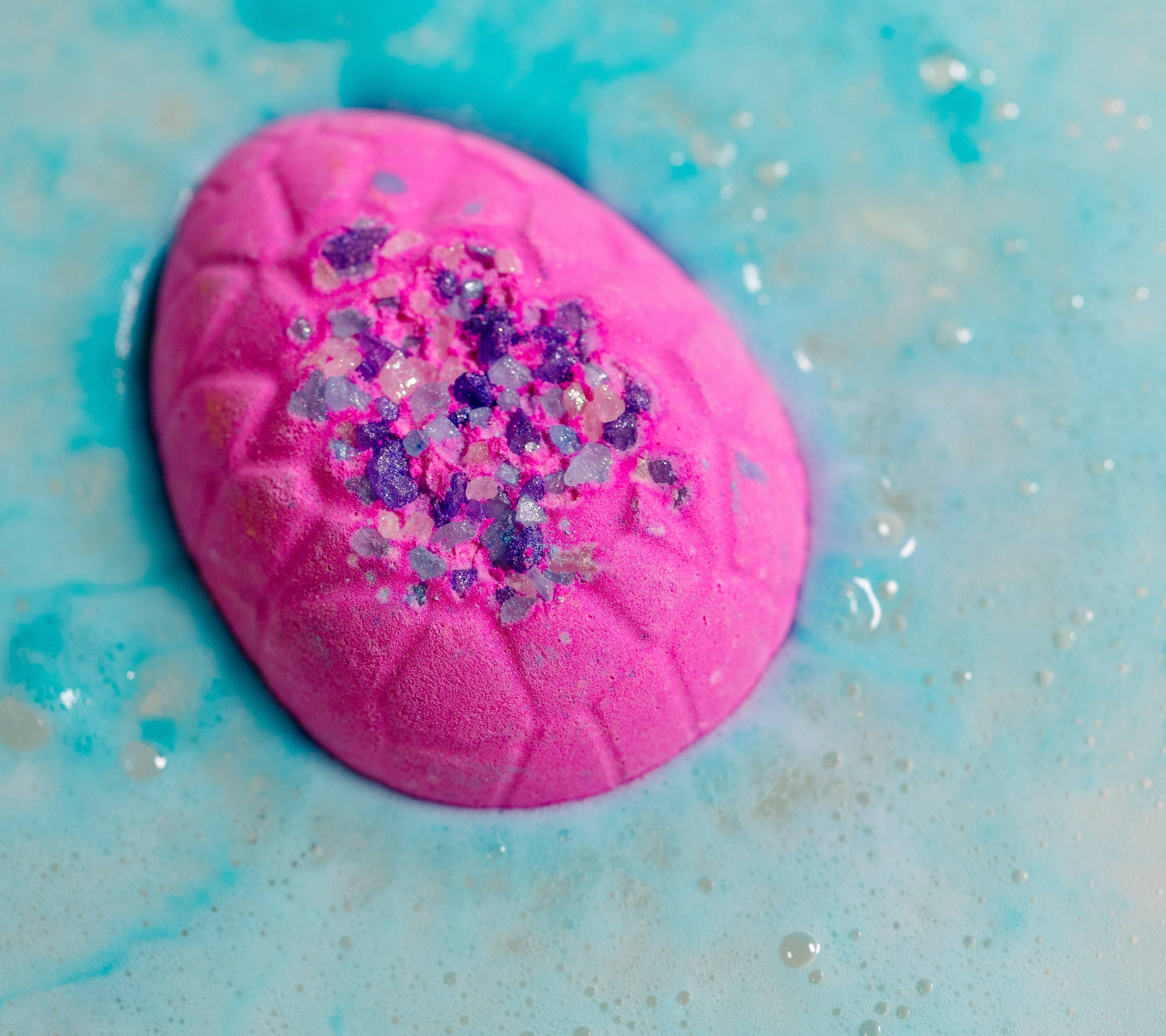 The bath bomb floats pink side up in baby blue coloured fizzing water, showing off the purple coloured sea salt embedded. 