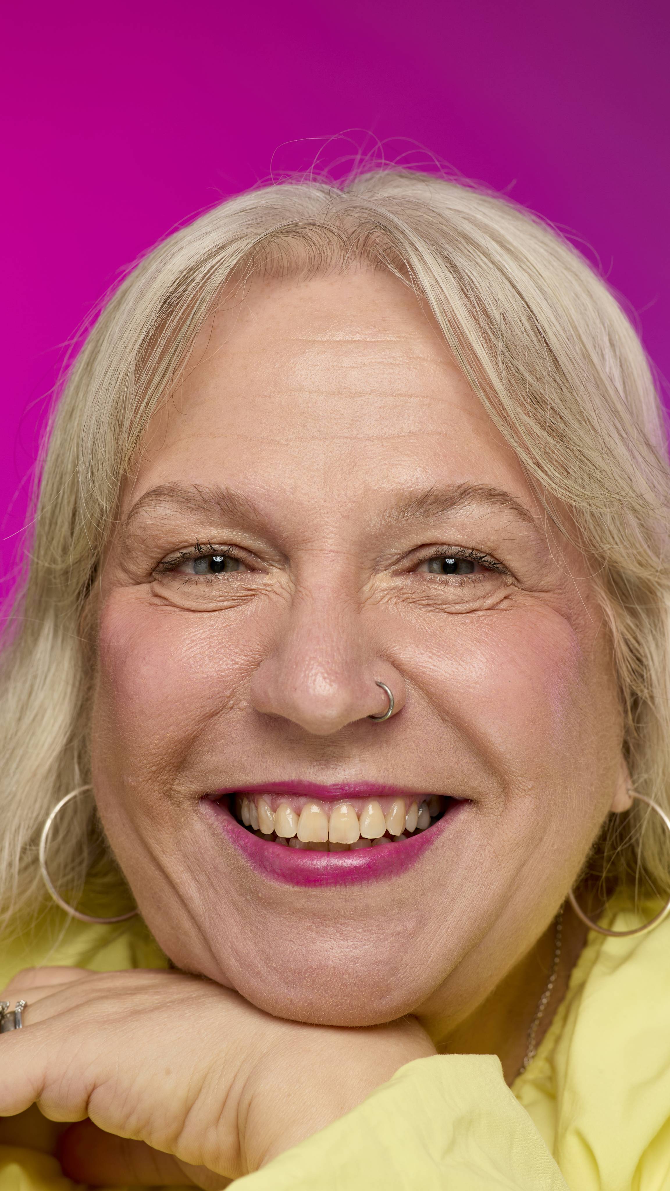 A smiling model with fair skin and light hair is shown on a vibrant pink background as they rest their chin on their hand. They smile, showing off the deep magenta Flash lipstick with a subtle shimmer. 