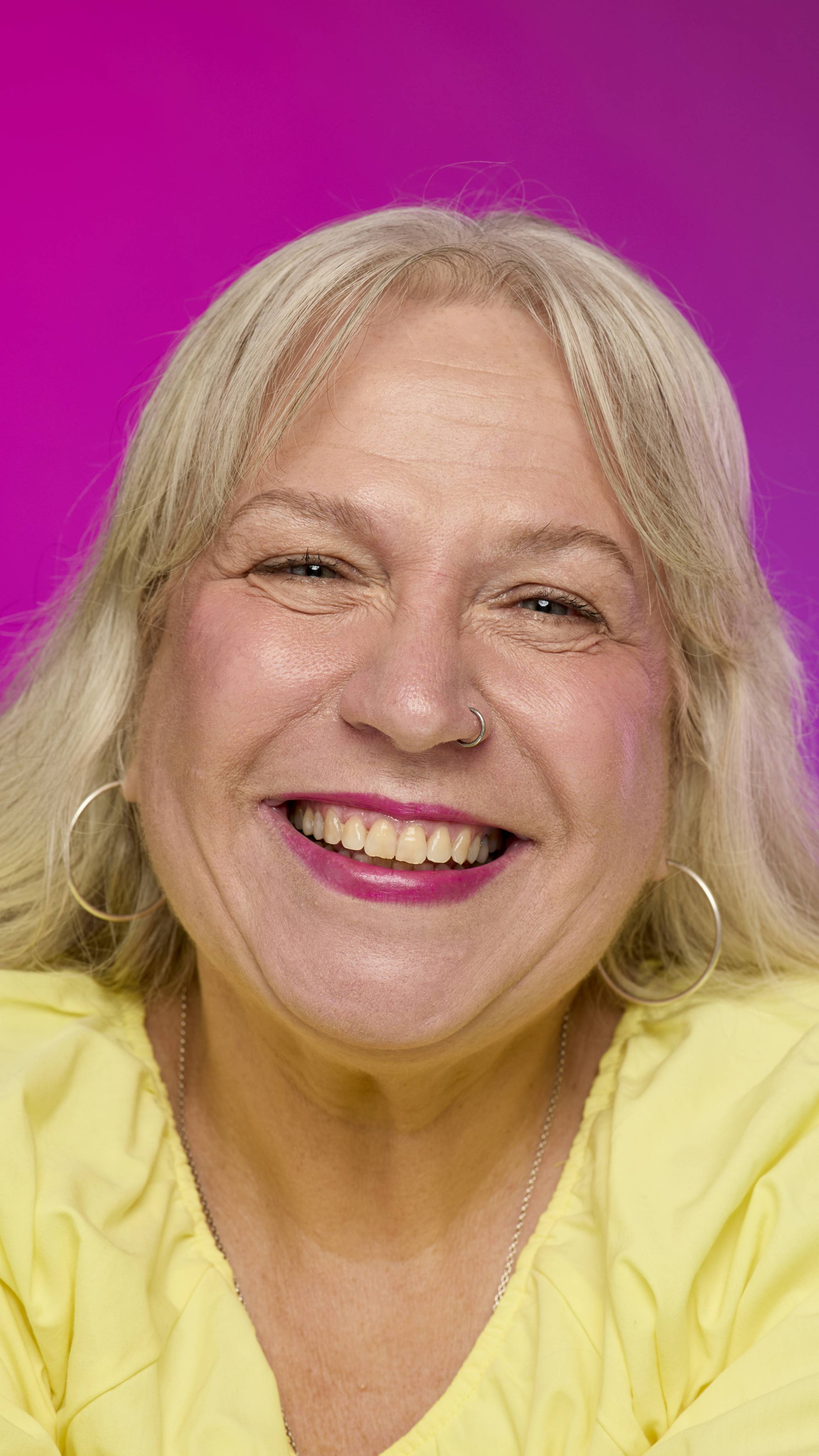 A smiling model with fair skin and light hair is shown on a vibrant pink background, wearing a bright yellow top. They smile, showing off the deep magenta Flash lipstick with a subtle shimmer. 