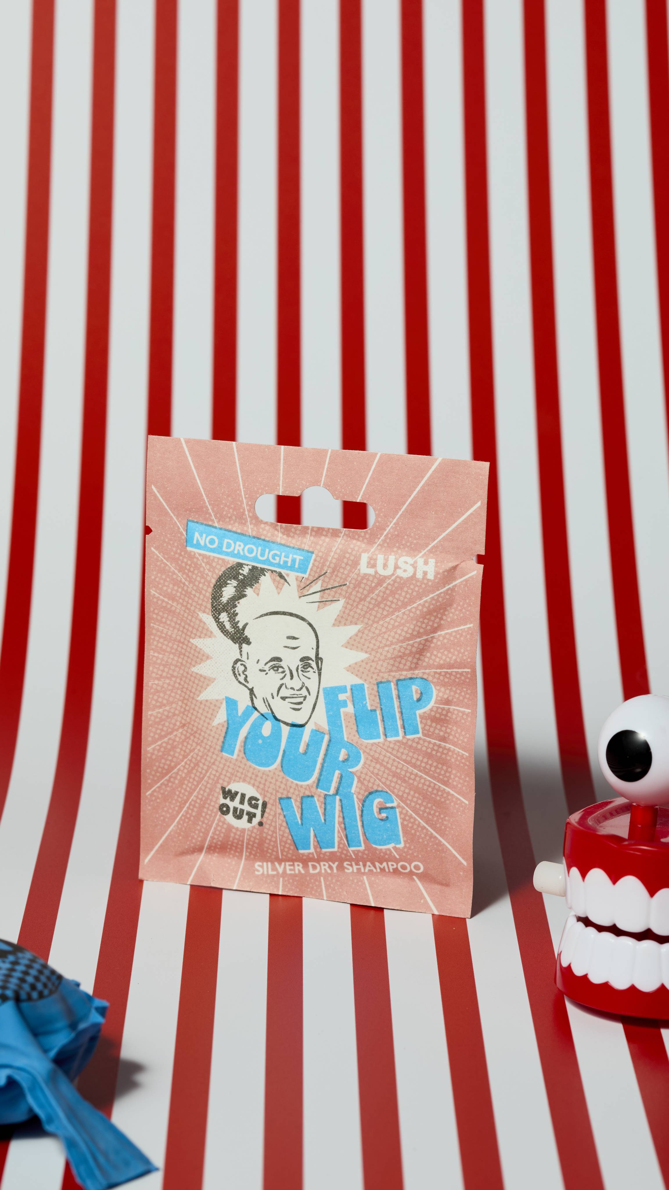 The image shows the Flip Your Wig, dry shampoo sachet product on a red and white, vertically striped background. There is a whoopie cushion and wind-up chattering teeth that are just visible.