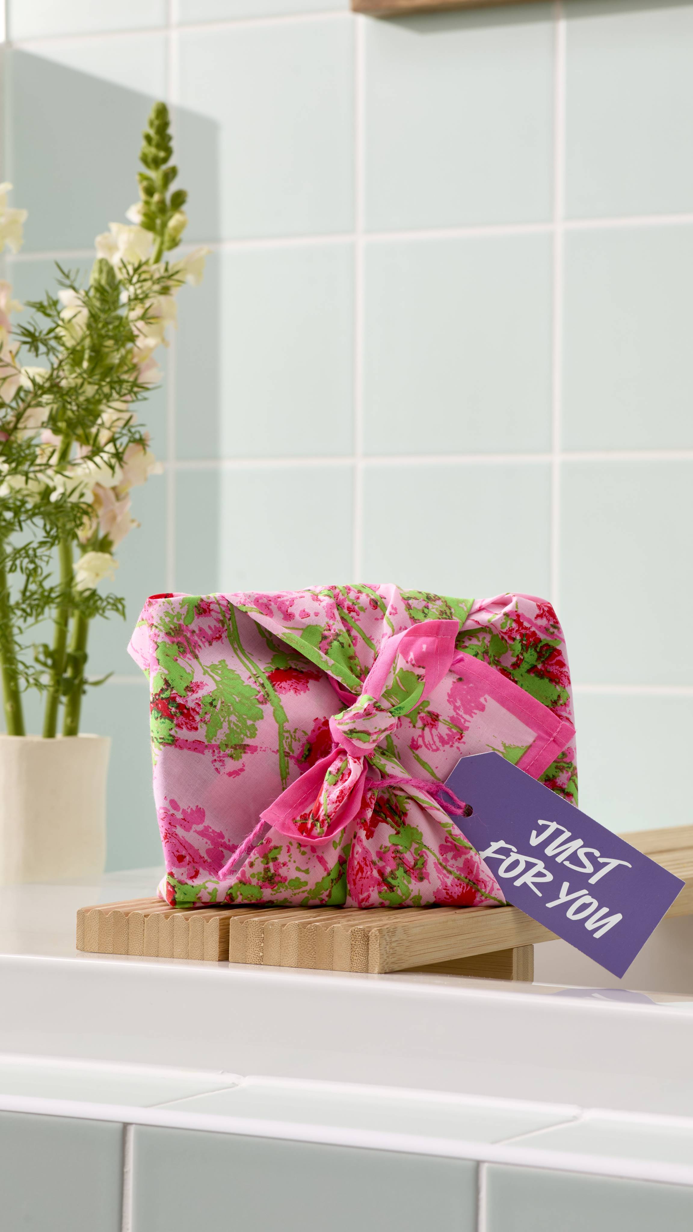 A gift wrapped in a "Floral Meadow" fabric knot-wrap sits on a wooden bath bridge over a white tub. The wrap features an impressionist design of red flowers and green stems on a light pink background, finished with a lilac "Just For You" gift tag.