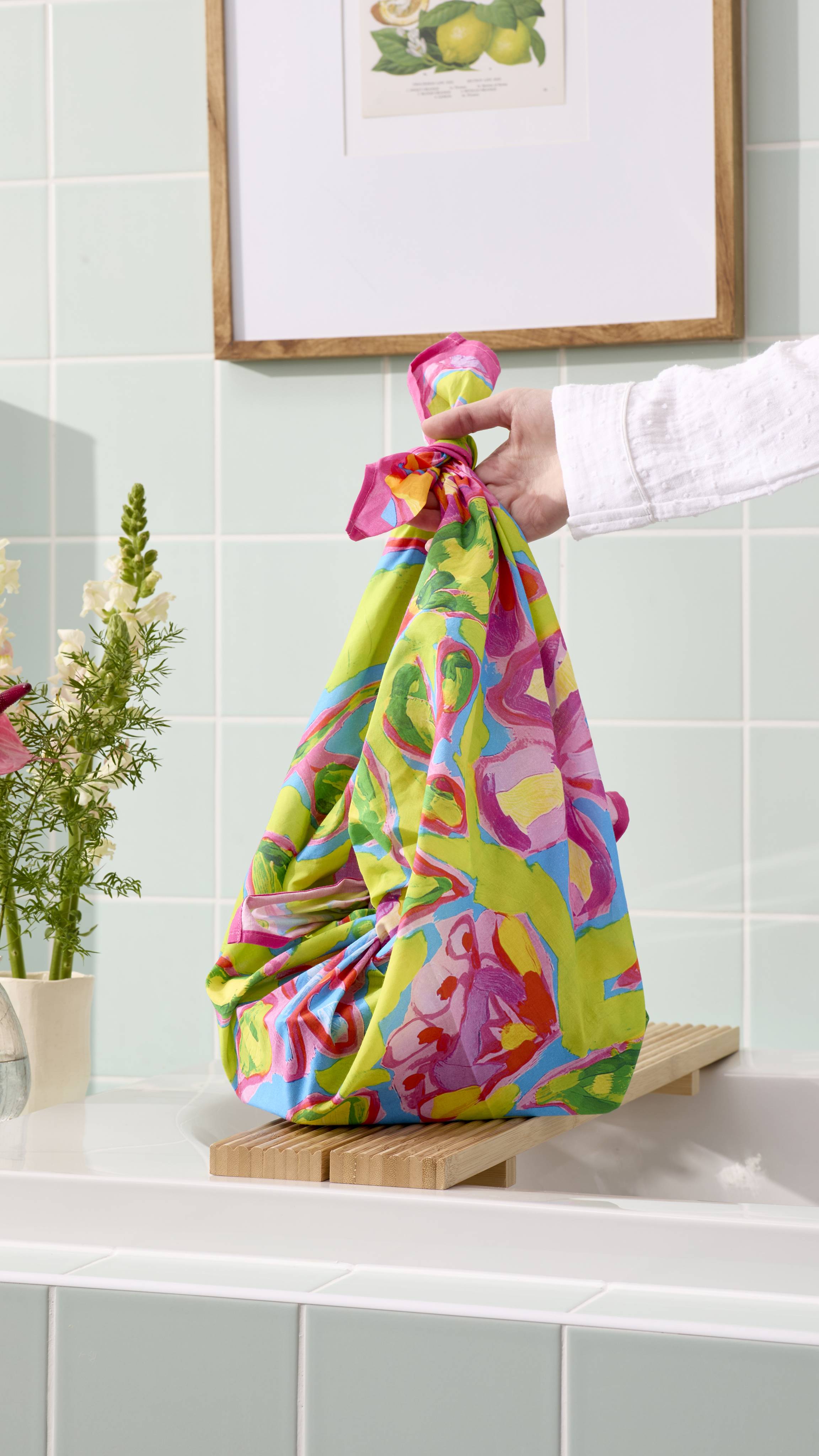 A person's hand holds the "Flourish In Colour" fabric knot-wrap, tied at the top to form a bag. The vibrant material features pink and blue flowers on a lime-green background. It rests on a wooden bath bridge in a bathroom with pale green tiles.