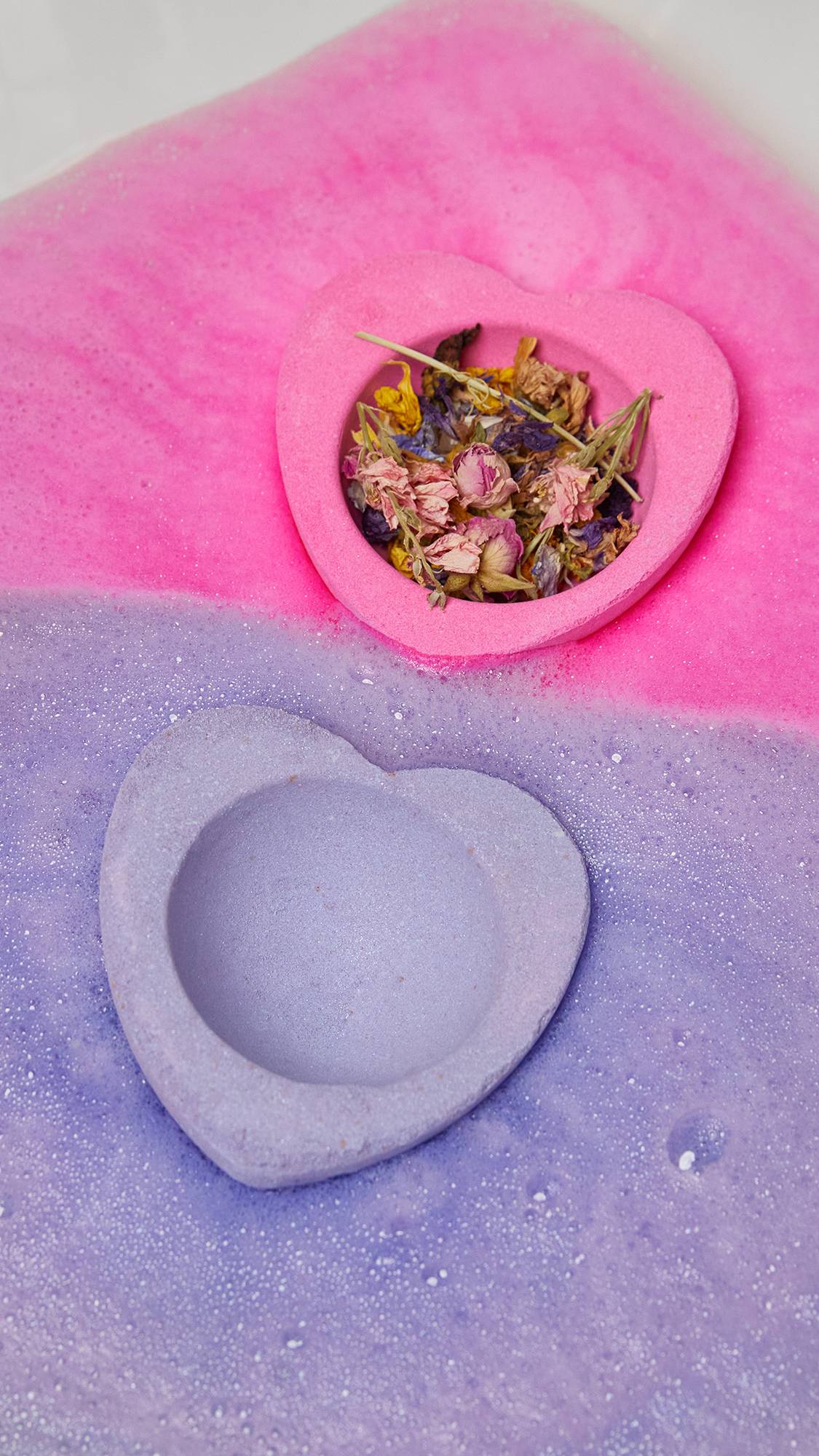Image shows the two halves of the bath bomb dissolving at the same time with a solid colour divide. The pink side holds dried flowers. 