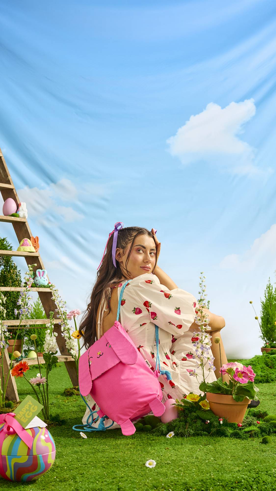 The model is sitting, next to a wooden ladder, in a white dress with strawberry decoration and they are wearing the Flower Bunny gift pink backpack over one shoulder.