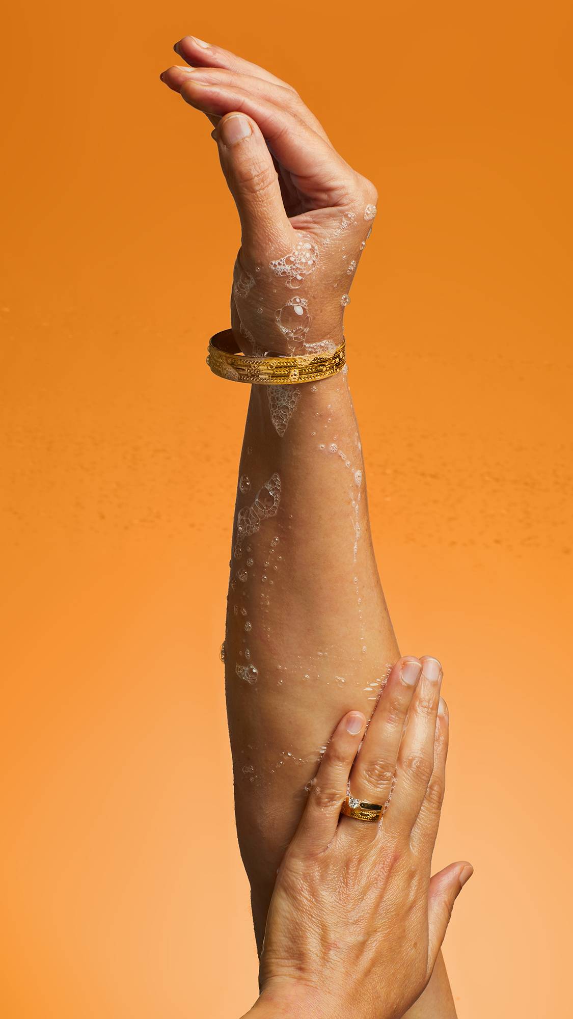 A close-up of the model's arm straight up on an orange background as they lather up soapy, Flower Gel suds. 