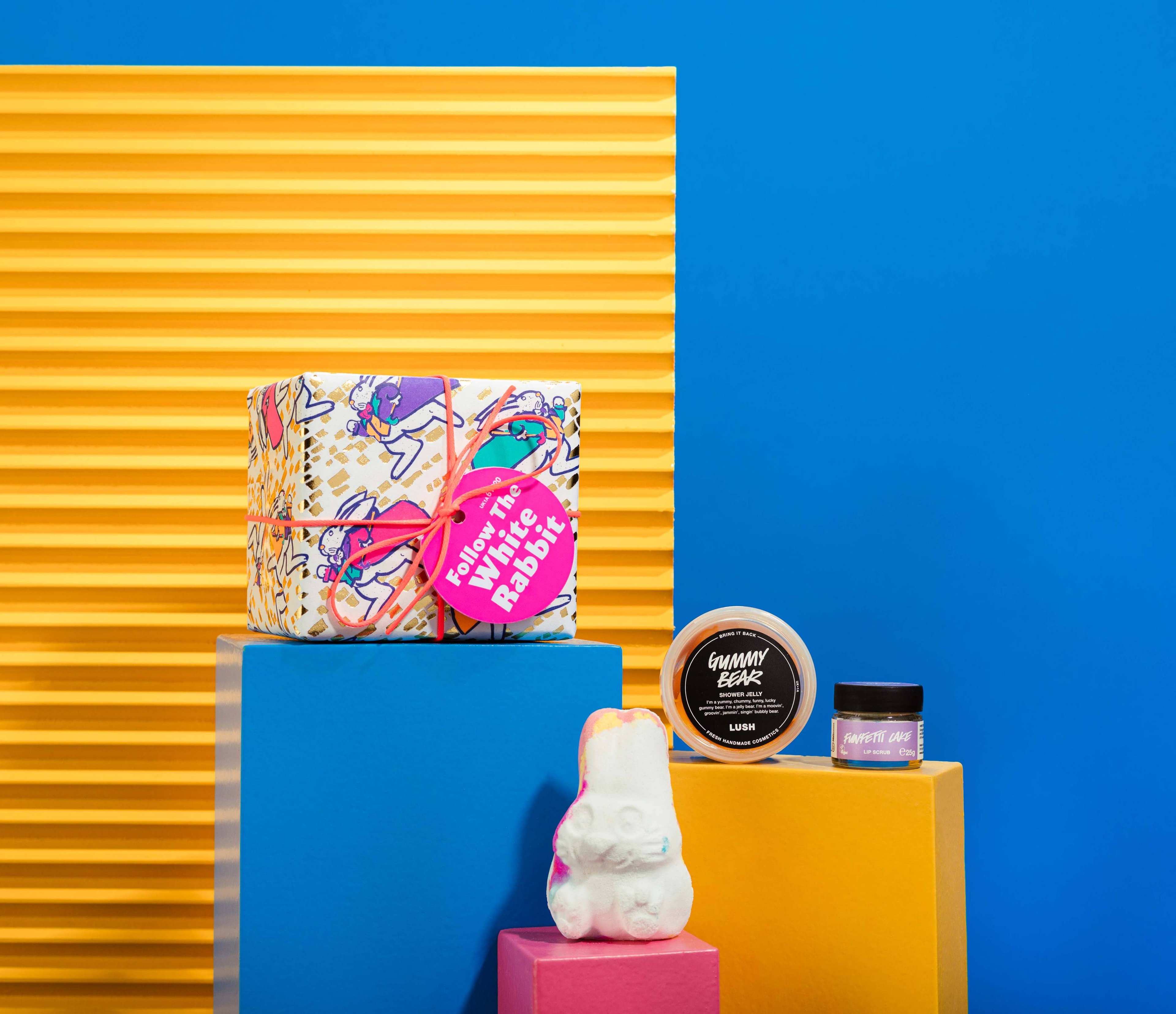 The gift in front of a blue and yellow background, surrounded by its products on blue, yellow and pink plinths.