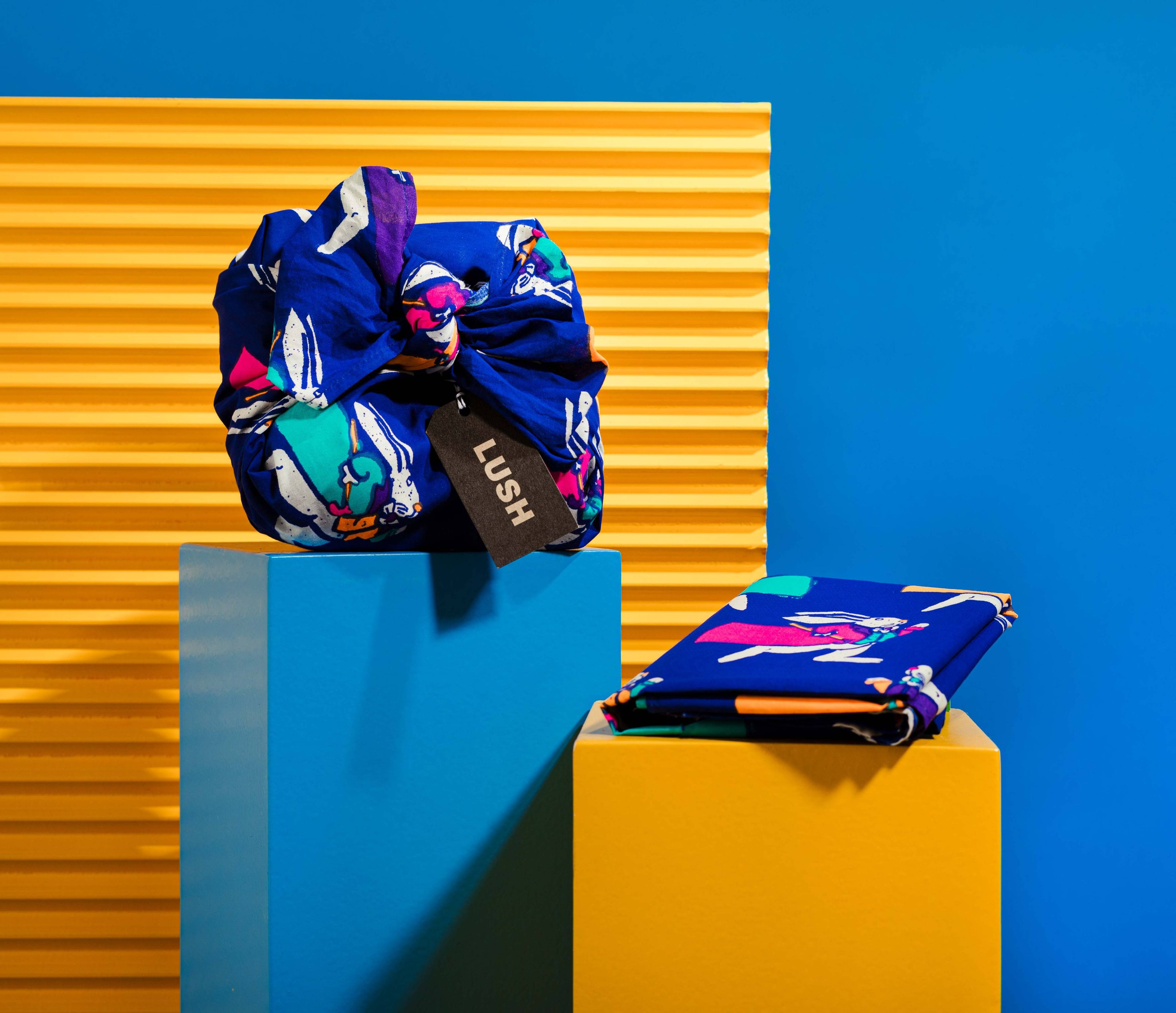 The Knot Wrap is decoratively wrapped with a Lush gift tag, alongside one folded, in front of a blue and yellow background. 