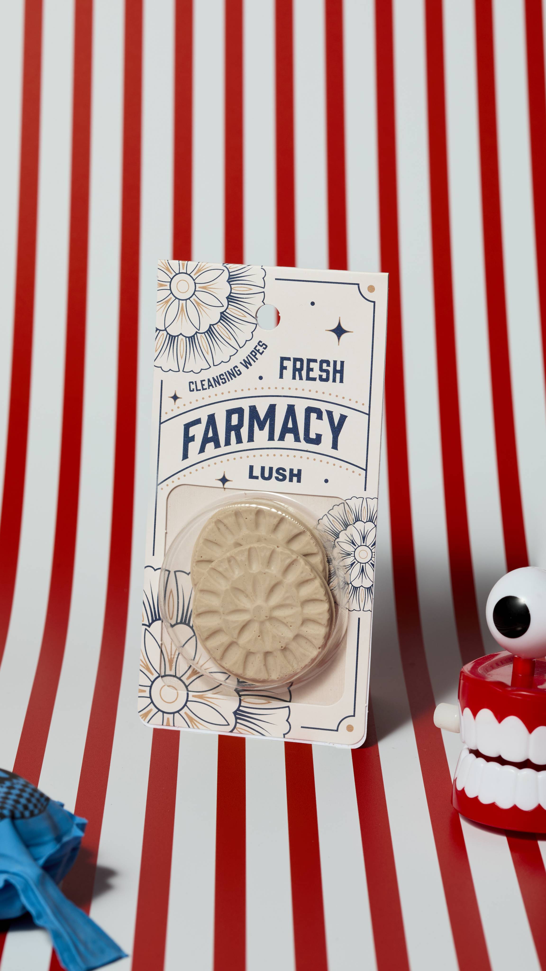 The image shows the Fresh Farmacy reusable cleansing pads product on a red and white, vertically striped background. There is a whoopie cushion and wind-up chattering teeth that are just visible.