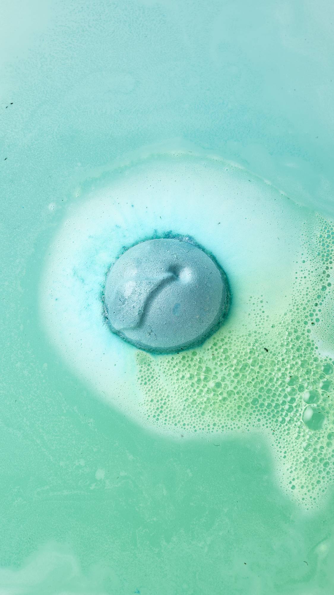 An aerial view shows the Freshly Ever After bath bomb fizzing in the water. The blue sphere is surrounded by a ring of bright white foam with splashes of vibrant aqua, sitting on water that has turned a light sea-green.