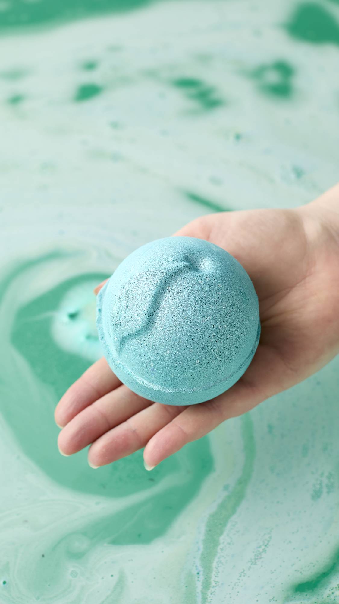 A person's hand holds a bright aqua-blue Freshly Ever After bath bomb over swirling bath water. The water beneath is a marbled pattern of foamy white and deep mint green.
