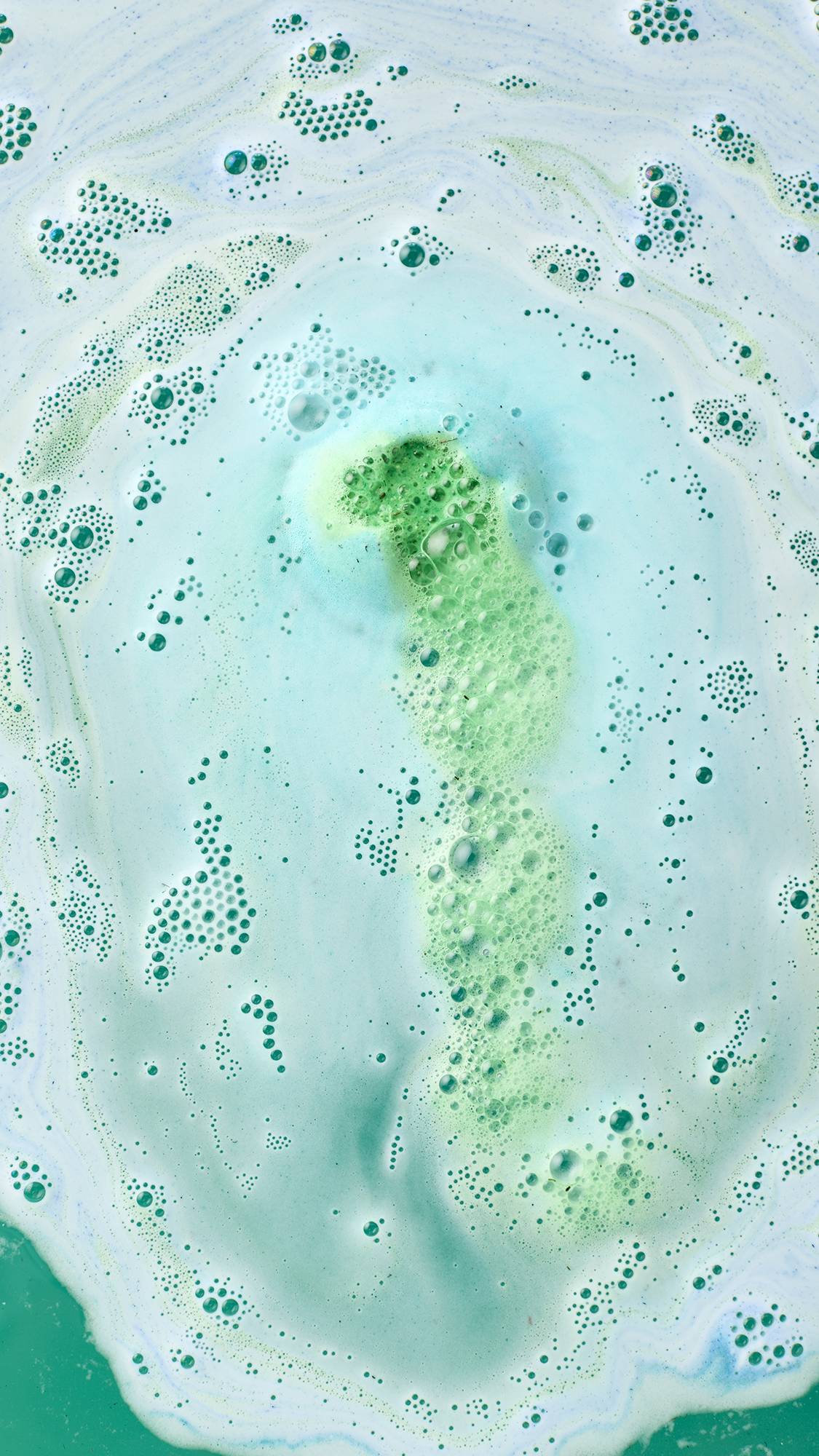 An aerial close-up of the Freshly Ever After bath bomb dissolving. The water's surface is a thick swirl of white and pale aqua foam, through which a vertical stream of vibrant lime green bubbles and foam is rising.