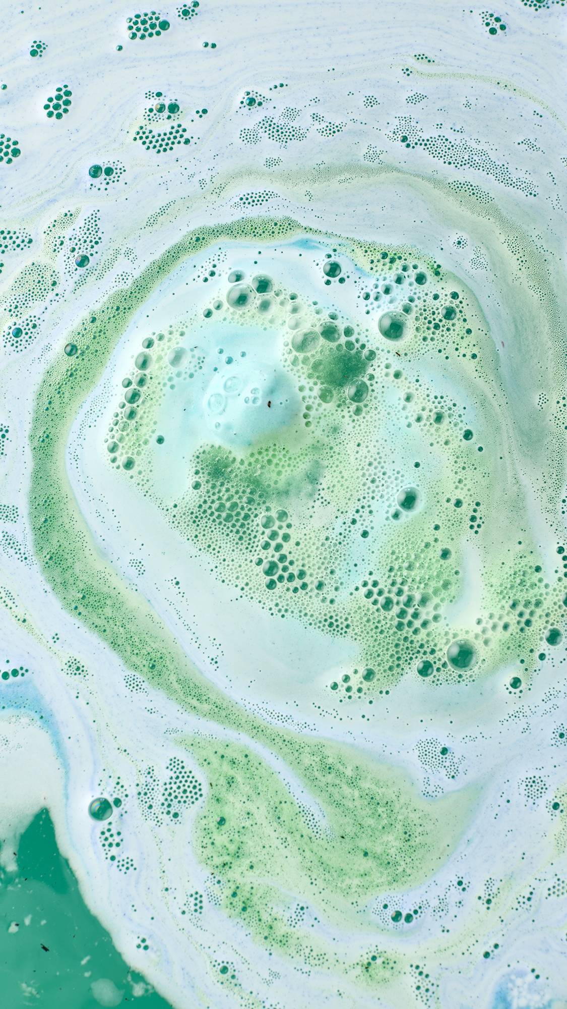 An aerial view of the Freshly Ever After bath bomb fully dissolving. The bath surface is covered in a thick, swirling pattern of white foam, pale aqua, and vibrant sea-green bubbles concentrated in a circular, marbled design.