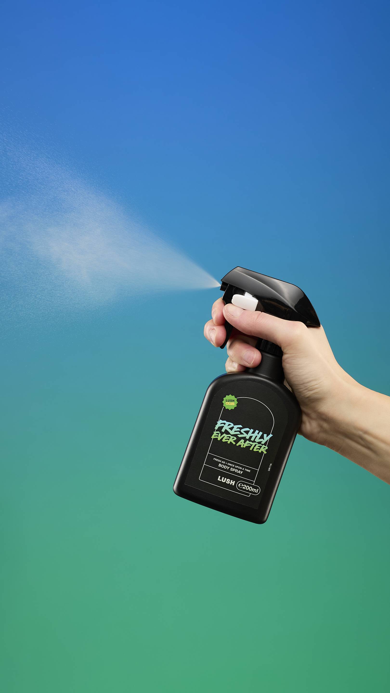 A hand holds the black, trigger-spray bottle of Freshly Ever After Body Spray, actively misting the spray to the left. The background is a vibrant gradient of deep blue at the top fading to bright green at the bottom.