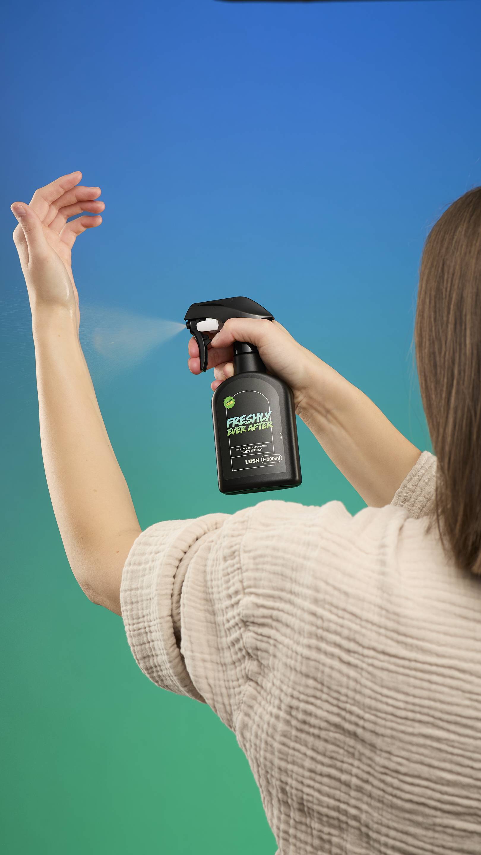 A person wearing a textured beige top is photographed from behind, holding the black Freshly Ever After Body Spray bottle and misting the product into the air above their head. The background is a vibrant blue-to-green gradient.
