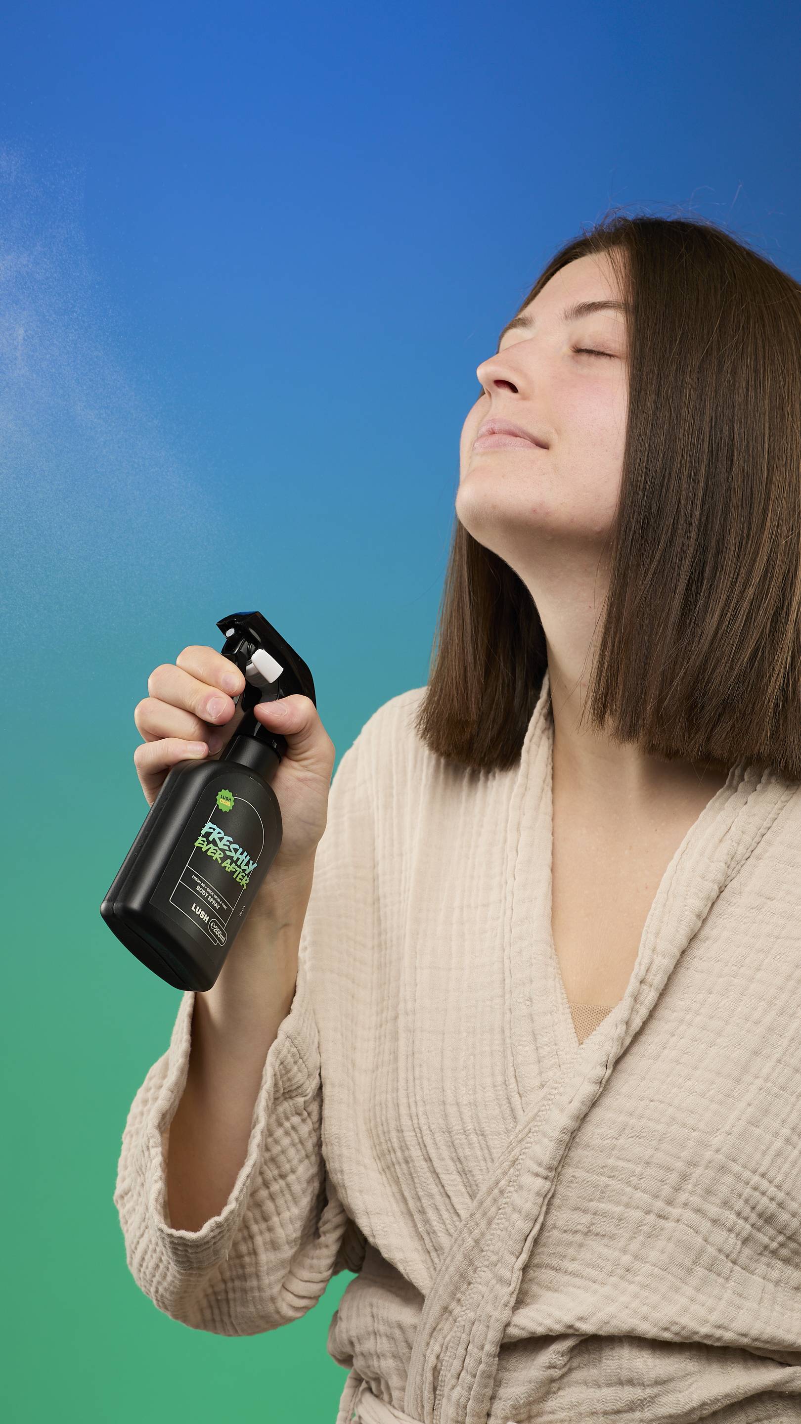 A smiling person wearing a beige robe holds the black Freshly Ever After Body Spray bottle and mists the product onto her neck and face. Her eyes are closed as she enjoys the spray, set against a blue-to-green gradient background.