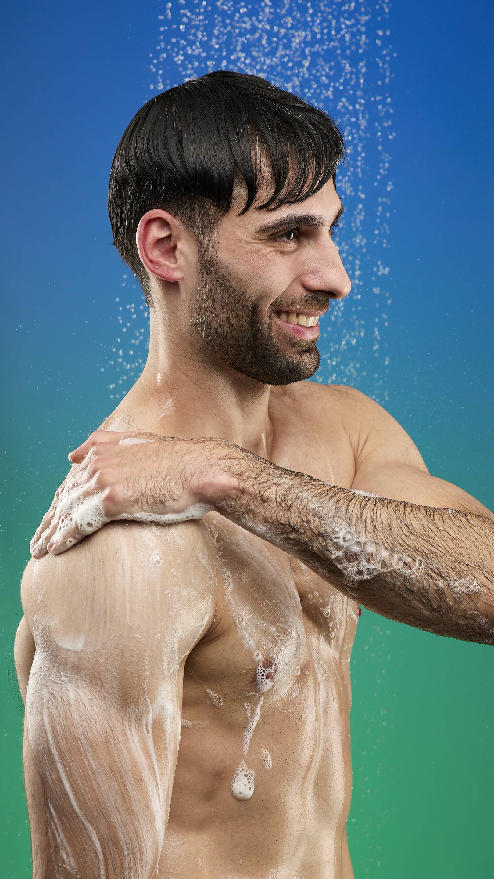 A close-up of a smiling man in the shower, facing right. He has lathered the Freshly Ever After shower gel into a white foam that covers his shoulder and chest as water sprays down from above. The background is a blue-to-green gradient.