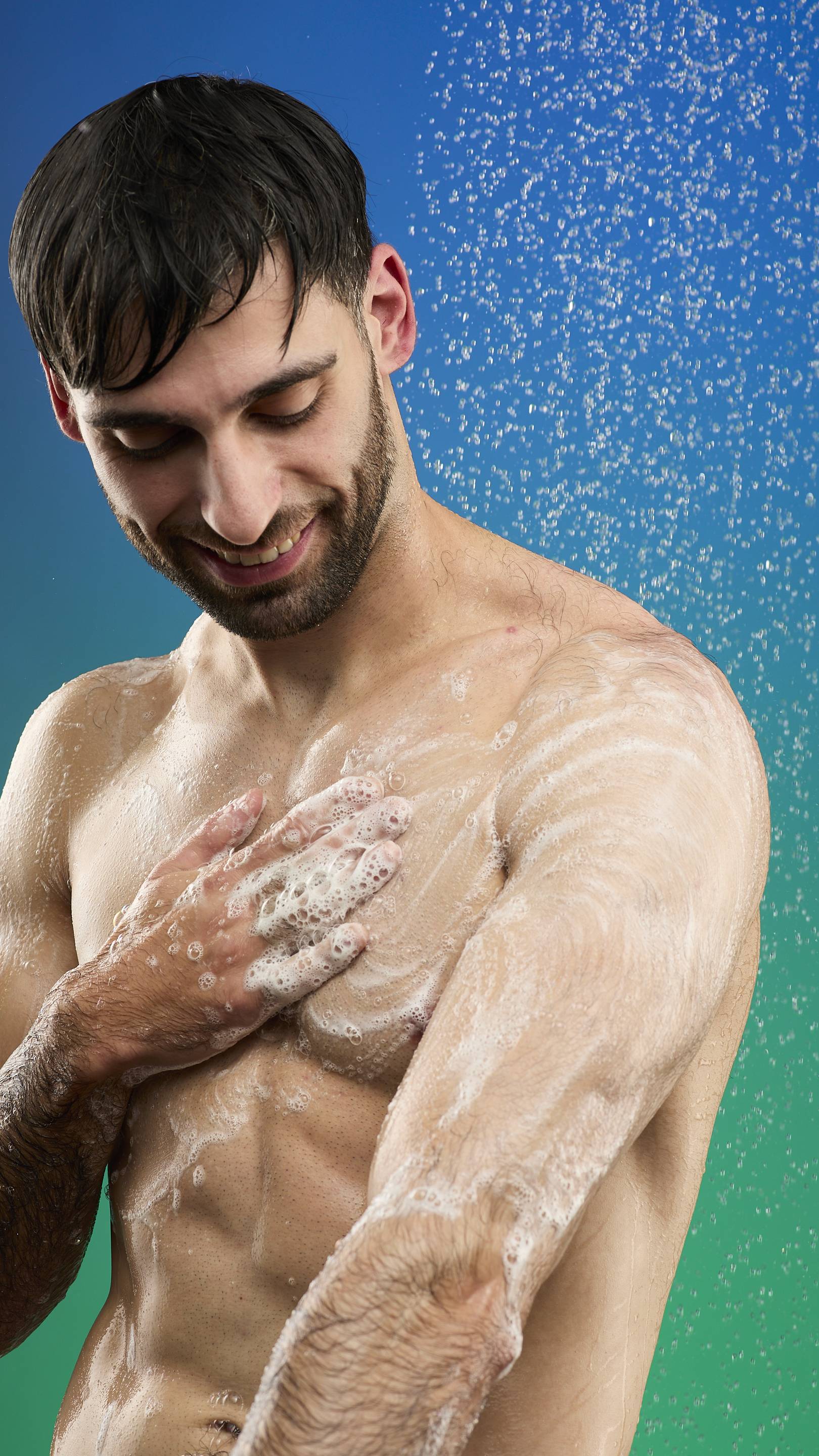 A bare-chested man smiles and looks down while lathering his arm and chest with the white foam of Freshly Ever After shower gel. The background is a blue and green shower gradient with water spraying over him.