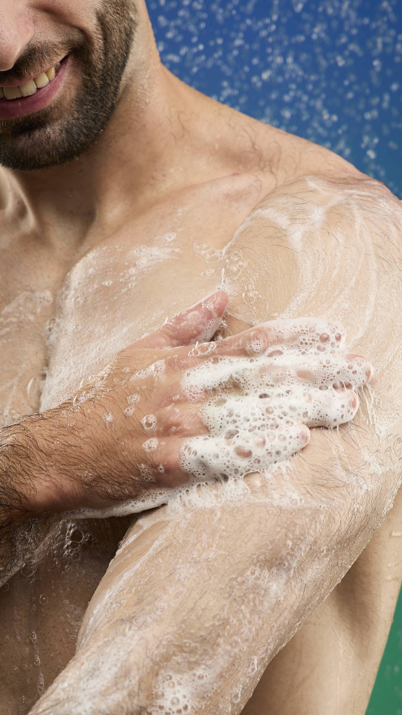 A close-up shot of a man's bare chest and arm, covered in the rich white lather from the Freshly Ever After shower gel. He is smiling slightly, with shower water visible in the blue background.