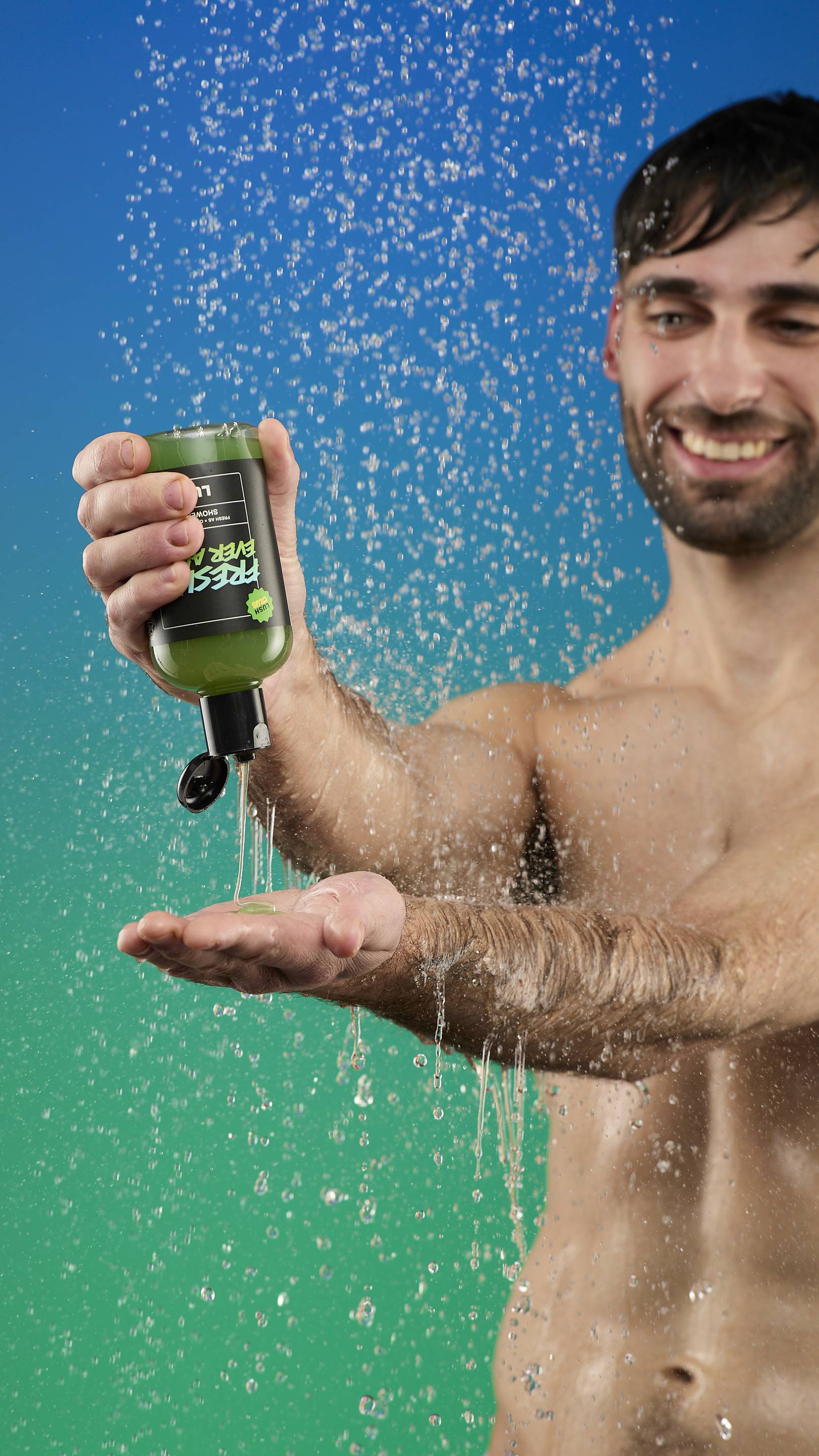 A smiling, bare-chested man is in a shower setting with a blue-to-green gradient background. He is pouring the Freshly Ever After shower gel (in its green-labeled bottle) onto his hand as water sprays down around him.