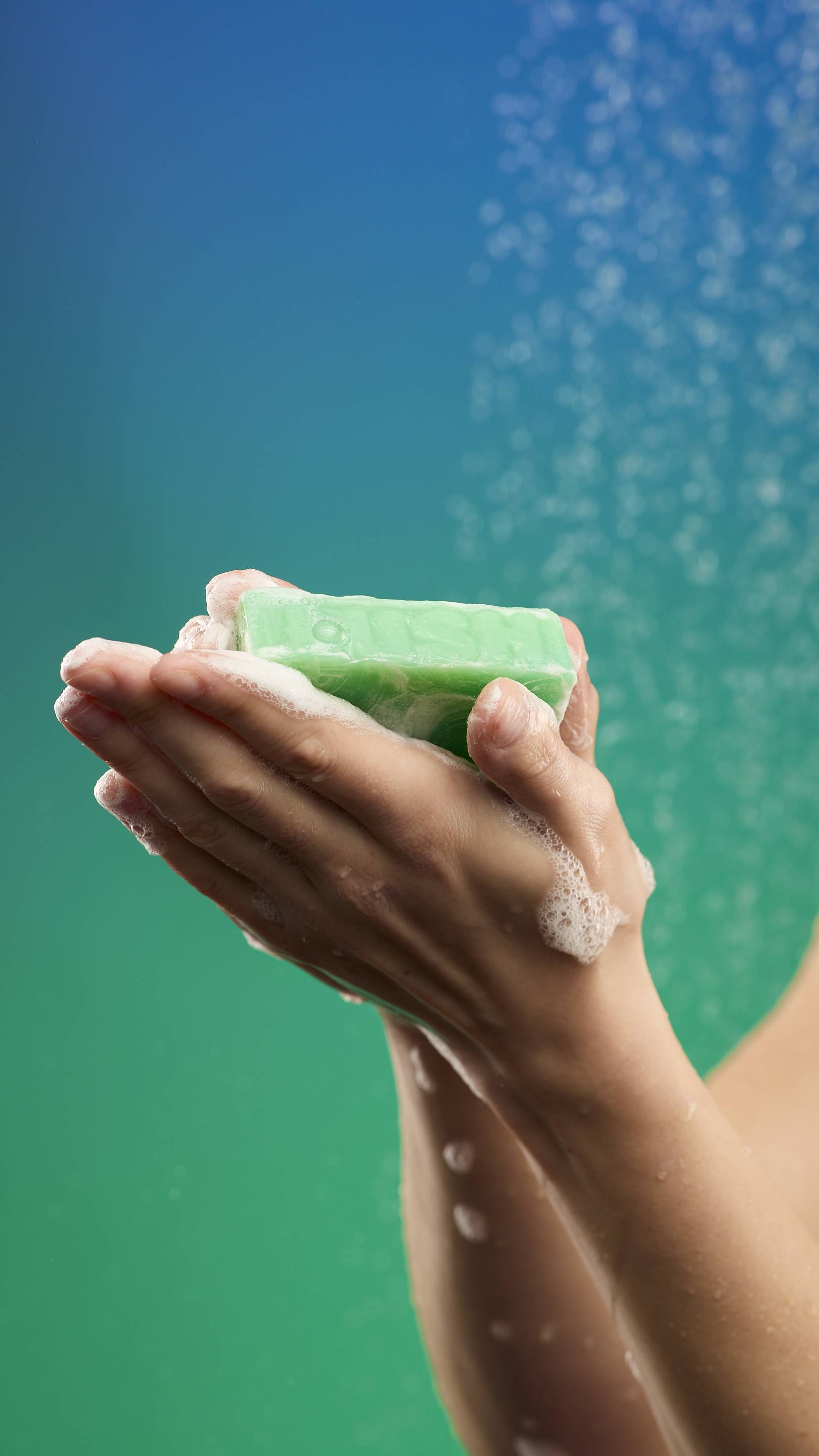 A pair of hands holds the marbled green Freshly Ever After soap bar, which is actively creating a small amount of white lather. The hands are held up against a shower background with a blue-to-green gradient and water droplets visible.