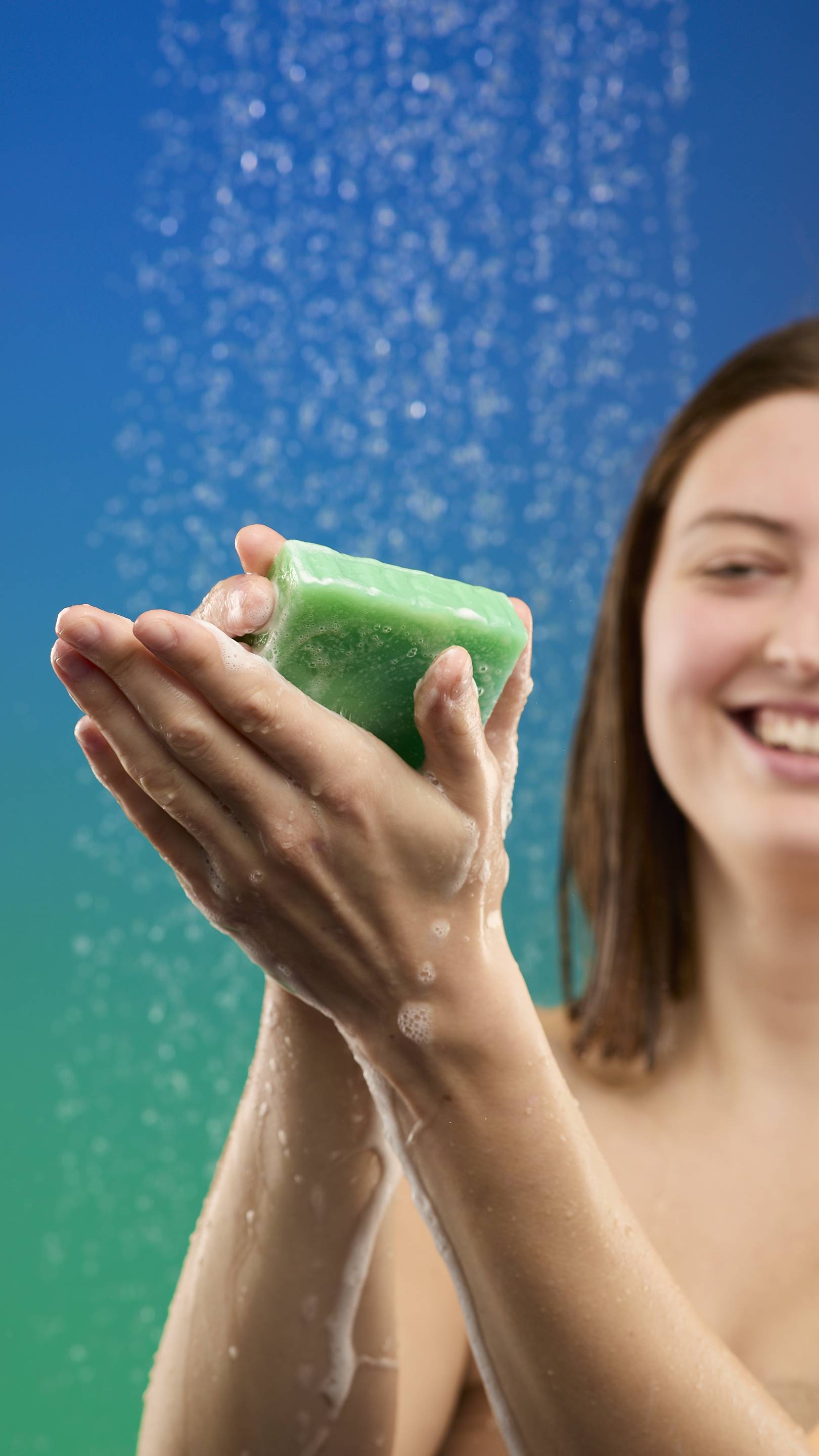 A smiling woman holds the large, marbled green Freshly Ever After soap bar in her hands, lathering it under a stream of shower water. The background is a bright blue-to-green gradient.