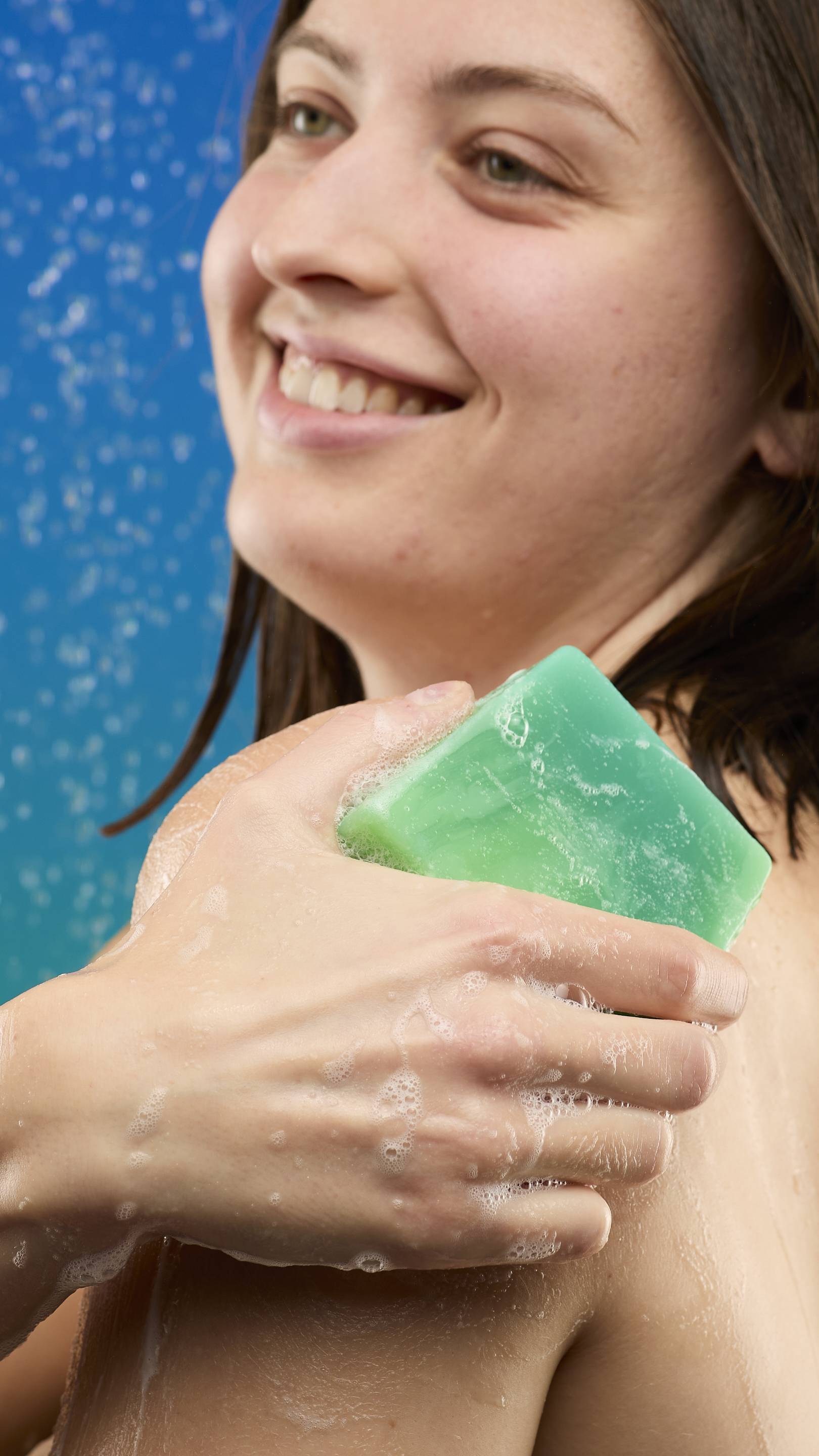 A close-up of a smiling woman in the shower, rubbing the marbled green Freshly Ever After soap bar across her shoulder. The soap is generating light white lather, and the background is a vivid blue.