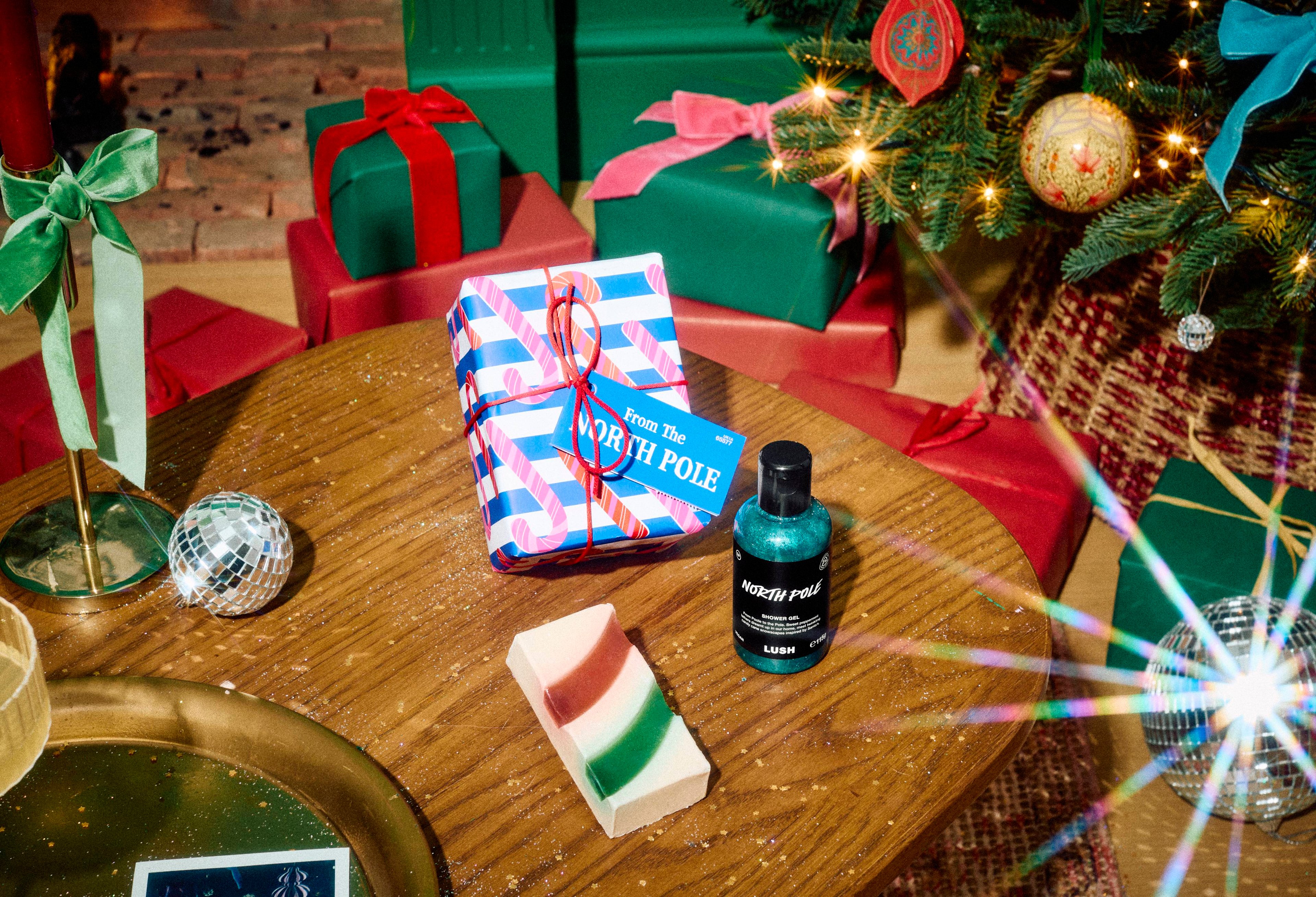 The Lush "From The North Pole" gift set sits on a wooden table beside a bottle of blue shower gel and a striped soap bar, in a festive room filled with wrapped presents and a decorated Christmas tree.
