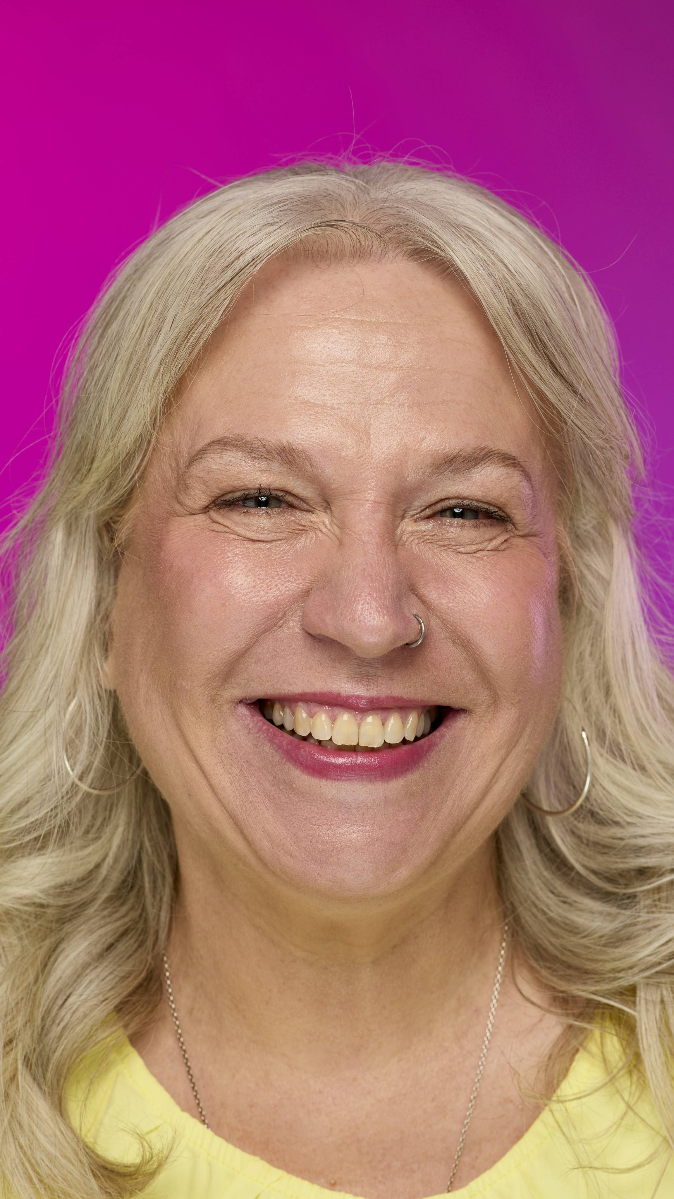 A smiling model with fair skin and light hair is shown from the neck up on a vibrant pink background. They smile, showing off the raspberry-red Fuse lipstick with a subtle shimmer. 
