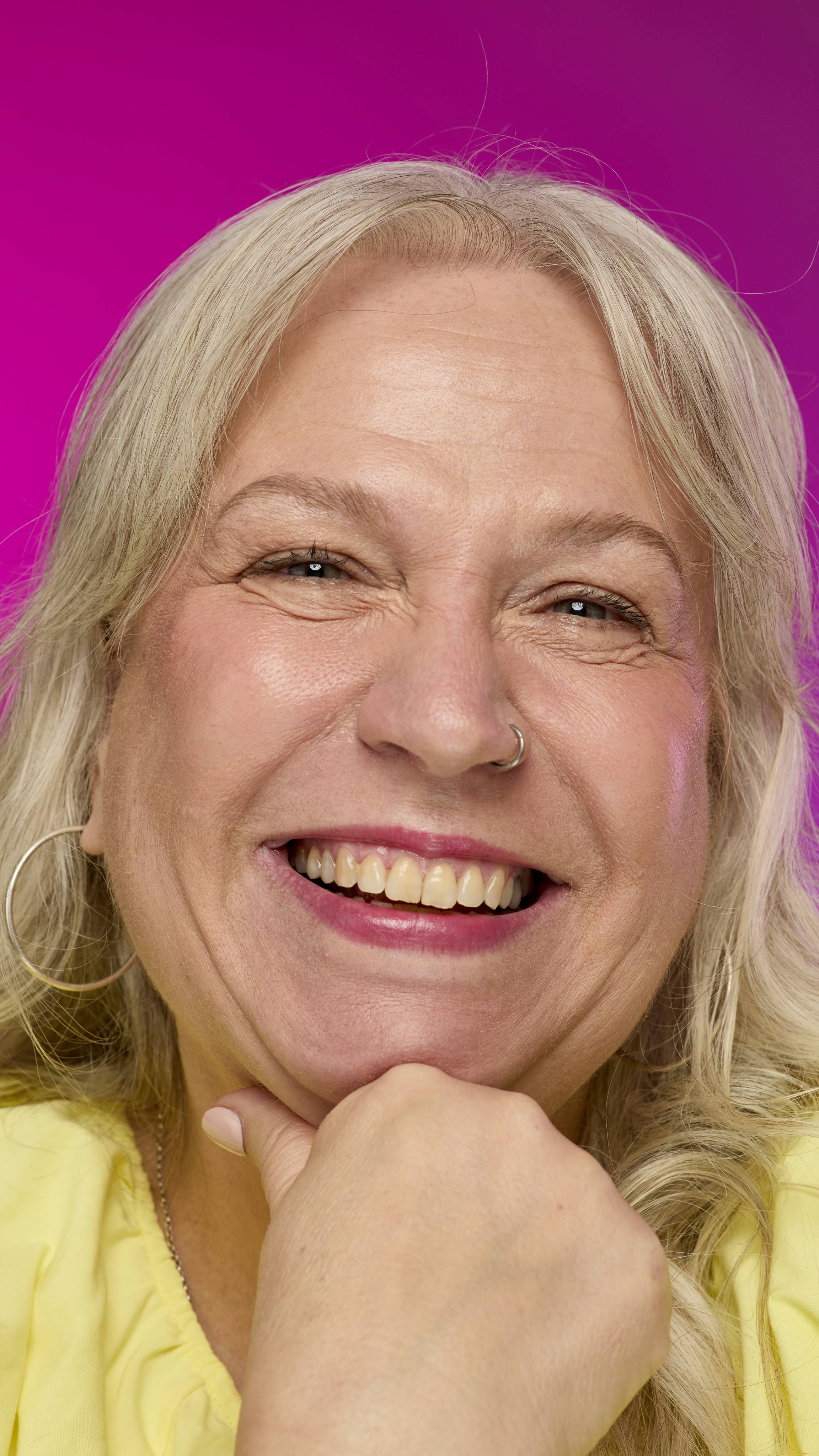 A smiling model with fair skin and light hair is shown from the neck up, with their chin resting on their hand, on a vibrant pink background. They smile, showing off the raspberry-red Fuse lipstick with a subtle shimmer. 