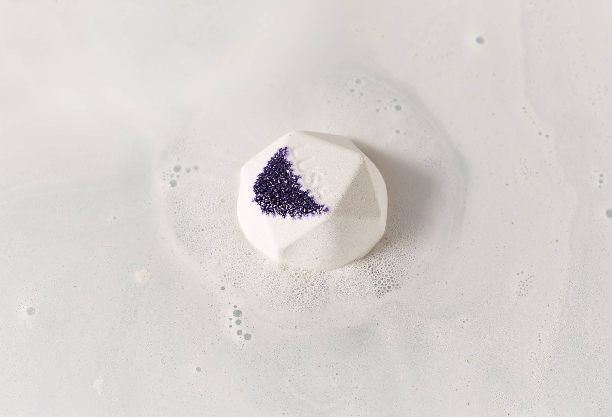 Fuurin bath bomb sits on calm, silky, white water with purple-coloured salt and "Lush" embossed on top. 