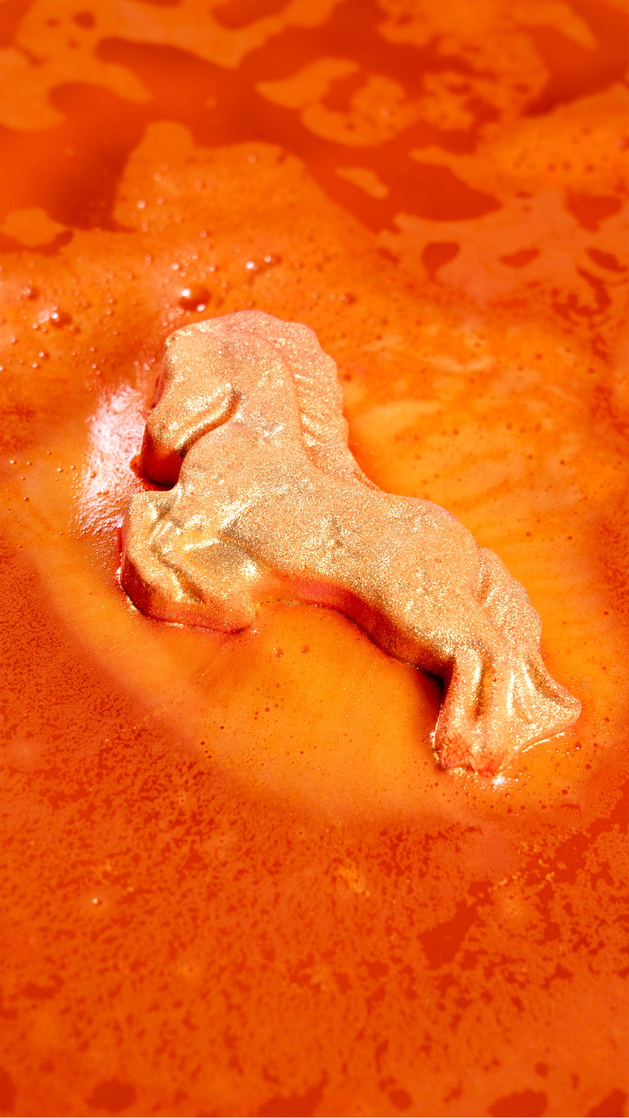 The gold, horse-shaped Gallop To Success Bath Bomb dissolves in bright orange water, releasing frothy foam and shimmering gold Plastic Free Glitter.