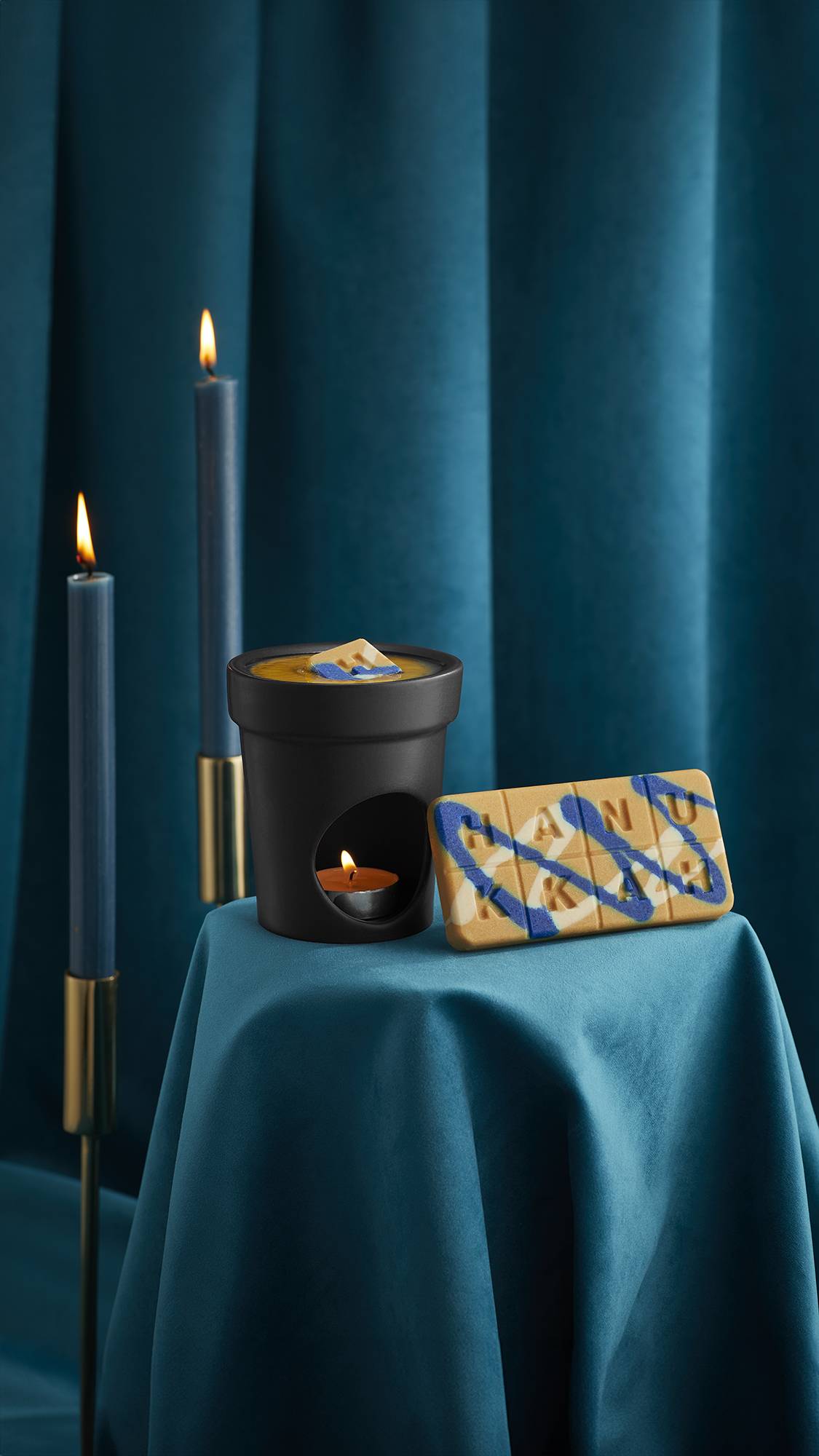 The Lush Gelt Melt is propped against a lit black wax burner with the melt on top, surrounded by deep teal velvet curtains.