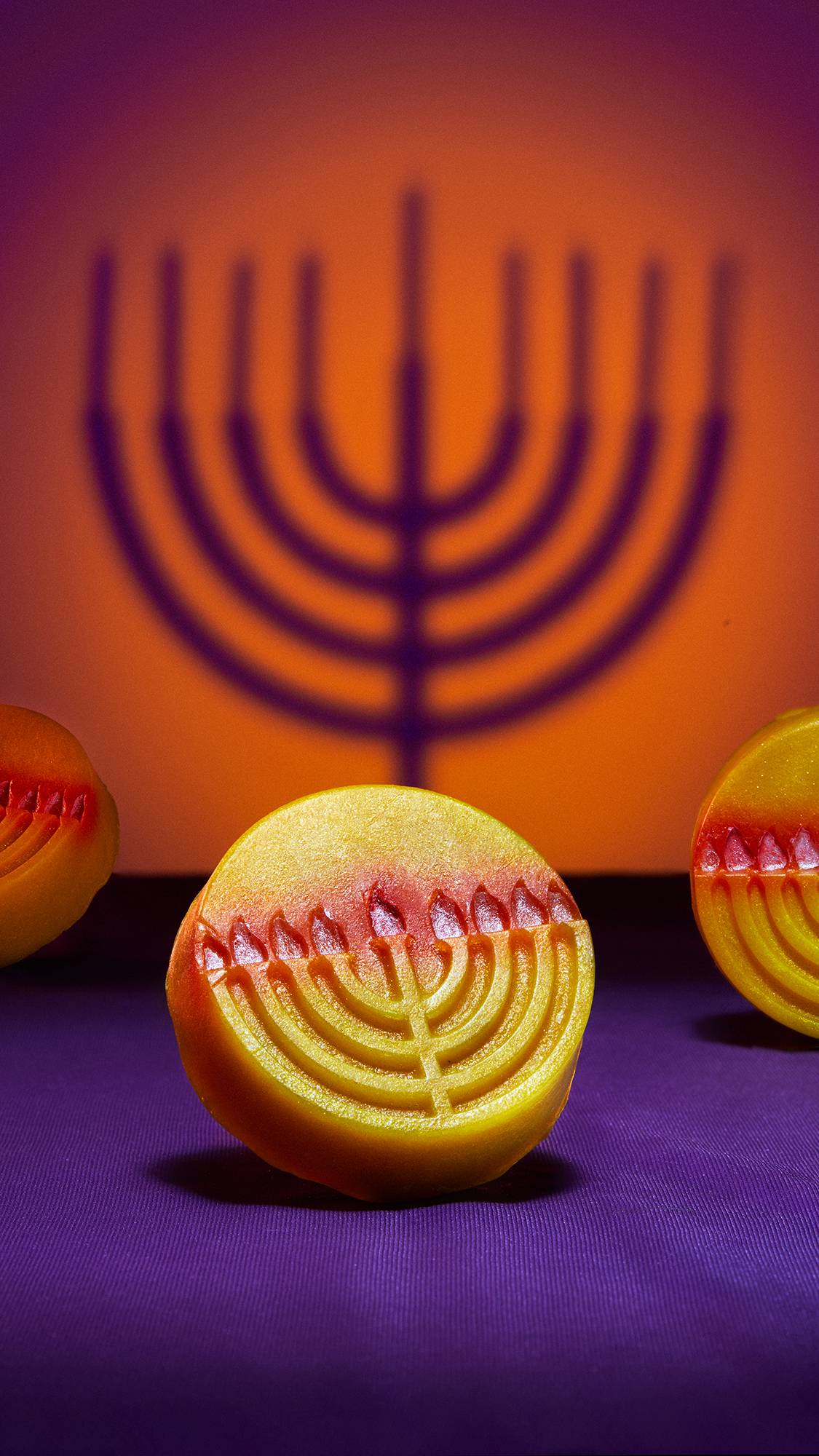 The image shows the Gelt soap propped at an angle focusing on the details of the embossed Menorah candelabra on the front. The background is a deep orange and purple colour with the shadow of a Menorah. 