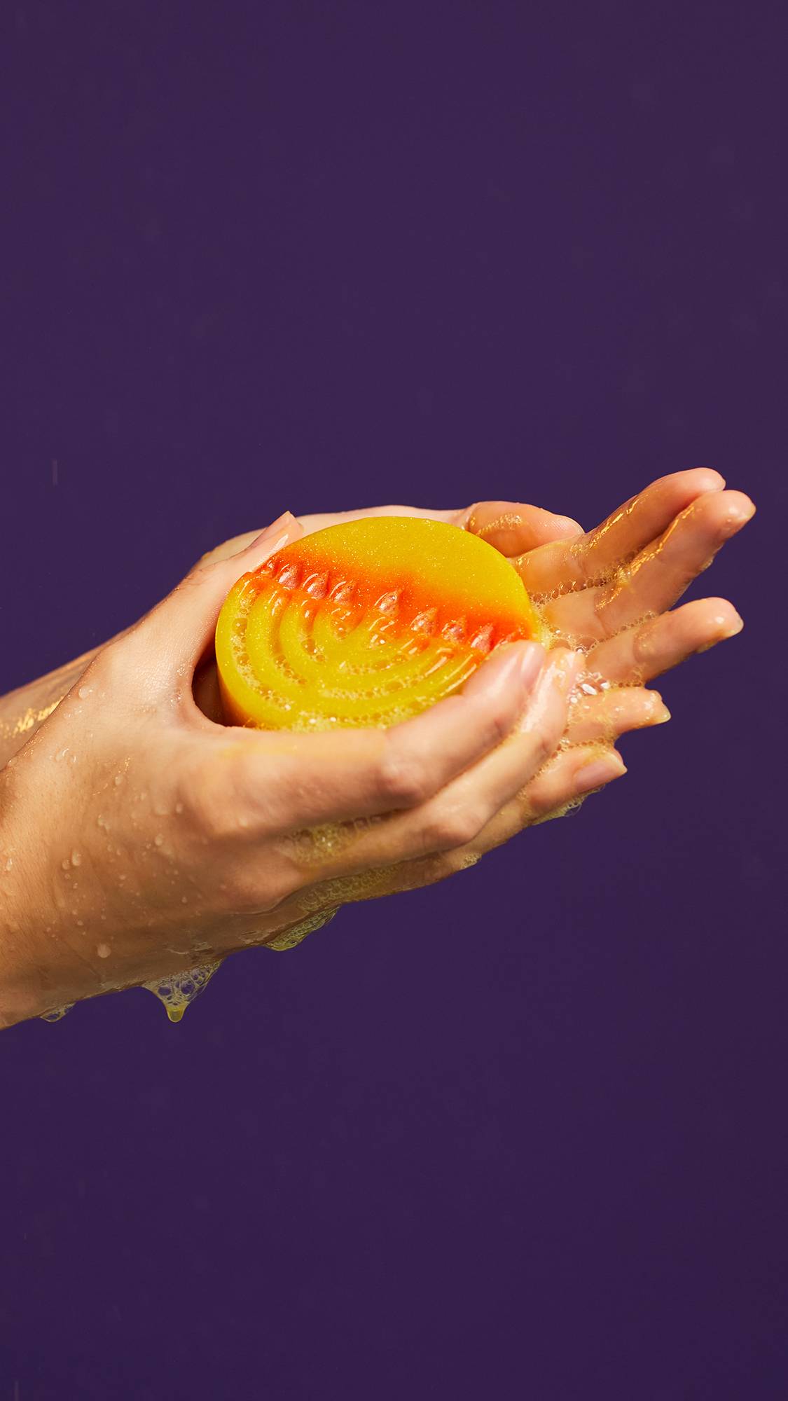 A close-up image of the model's hands gently holding the Gelt, Hanukkah-themed soap. Their hands are moist and they have lathered up delicate suds with the soap on a dark purple background.