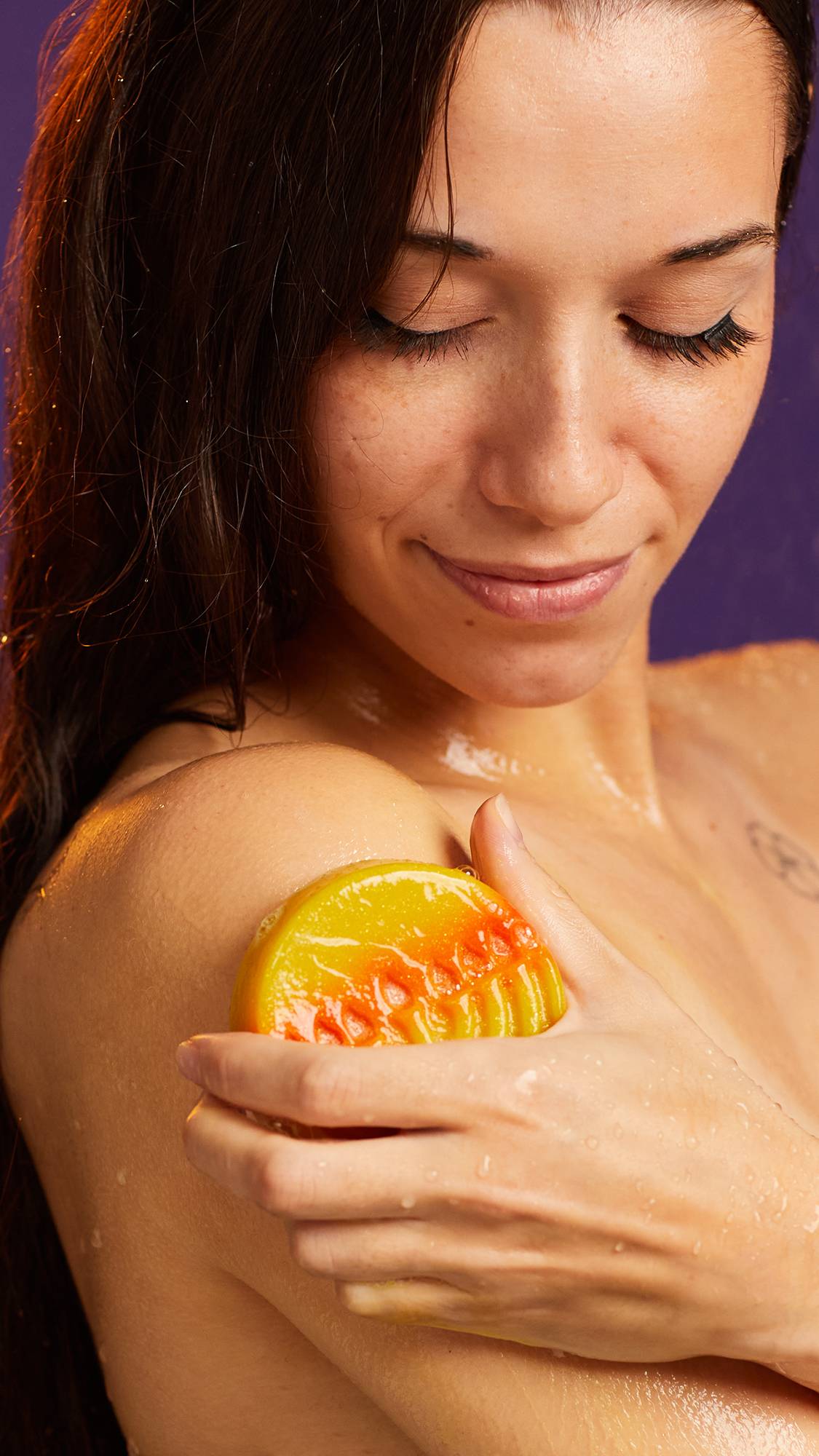 The image shows the model using the circular, Hanukkah-themed soap on their upper arm. They have dark, long hair that is down and their eyes are focused on the Gelt soap. 