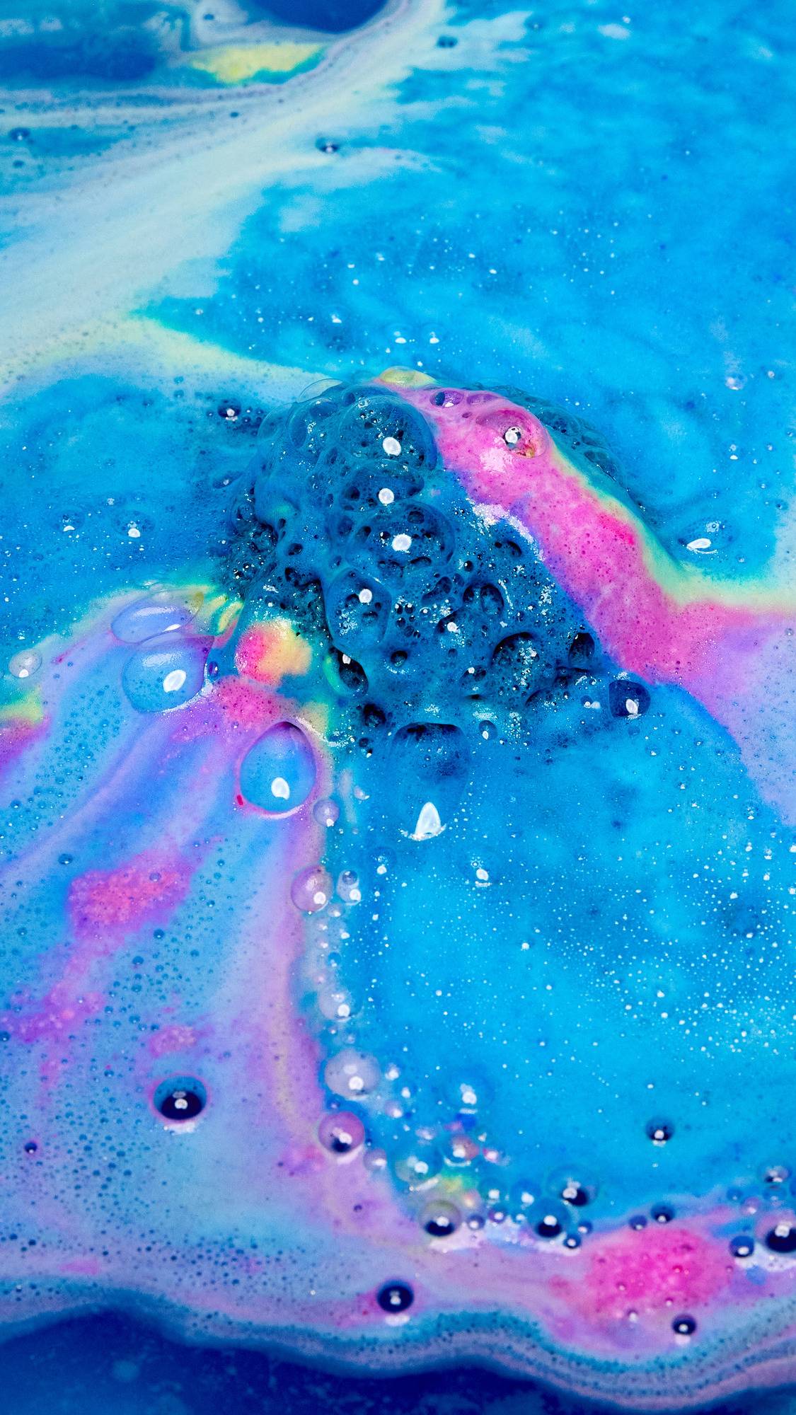 A close-up of the Giant Intergalactic bath bomb as it dissolves in the bath water releasing bright blue thick foam with flecks of pink and yellow. 