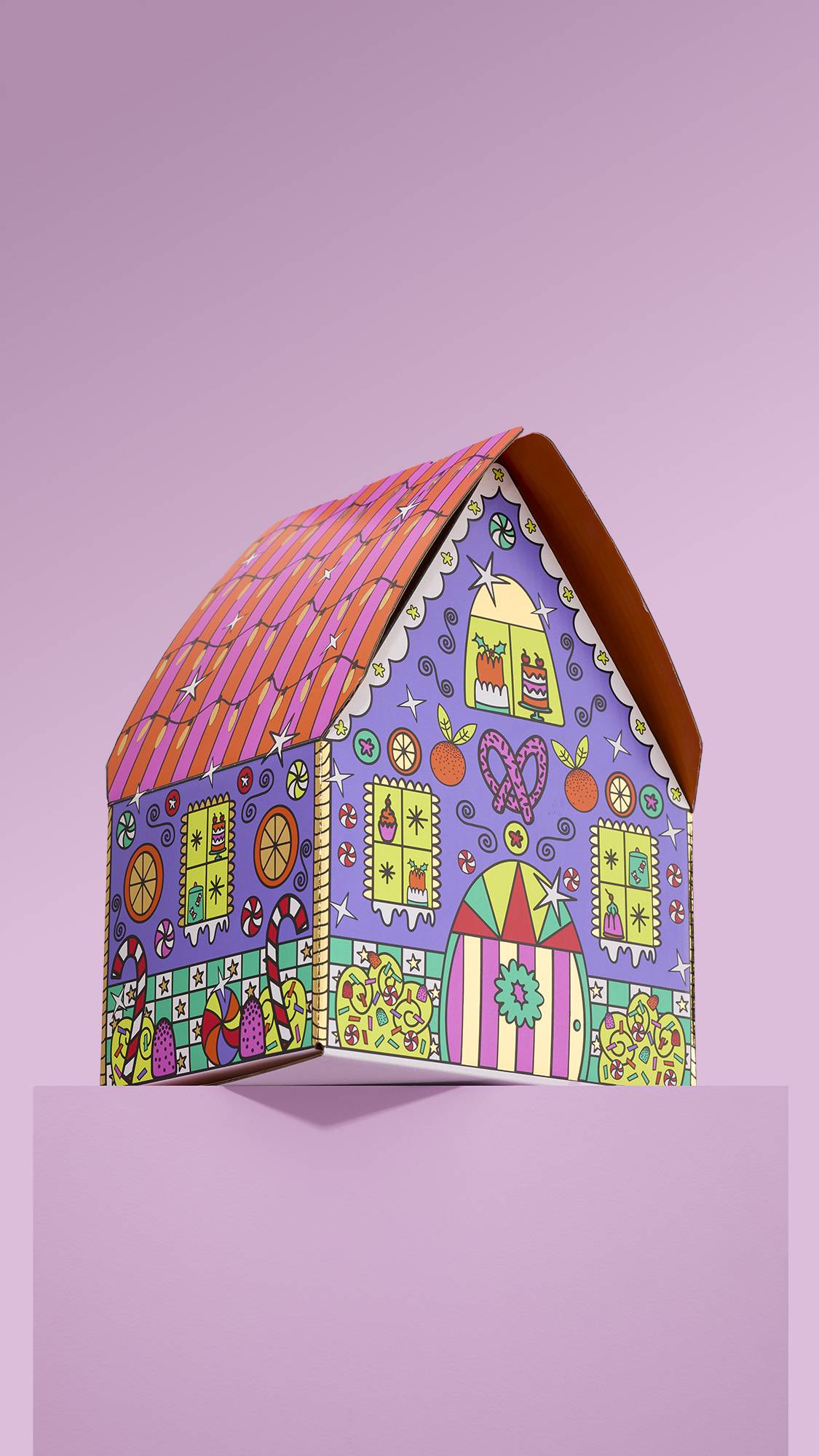 The image shows the vibrant,Gingerbread House gift box, with a colourful house design, sitting on a pink platform against a matching pink background. 