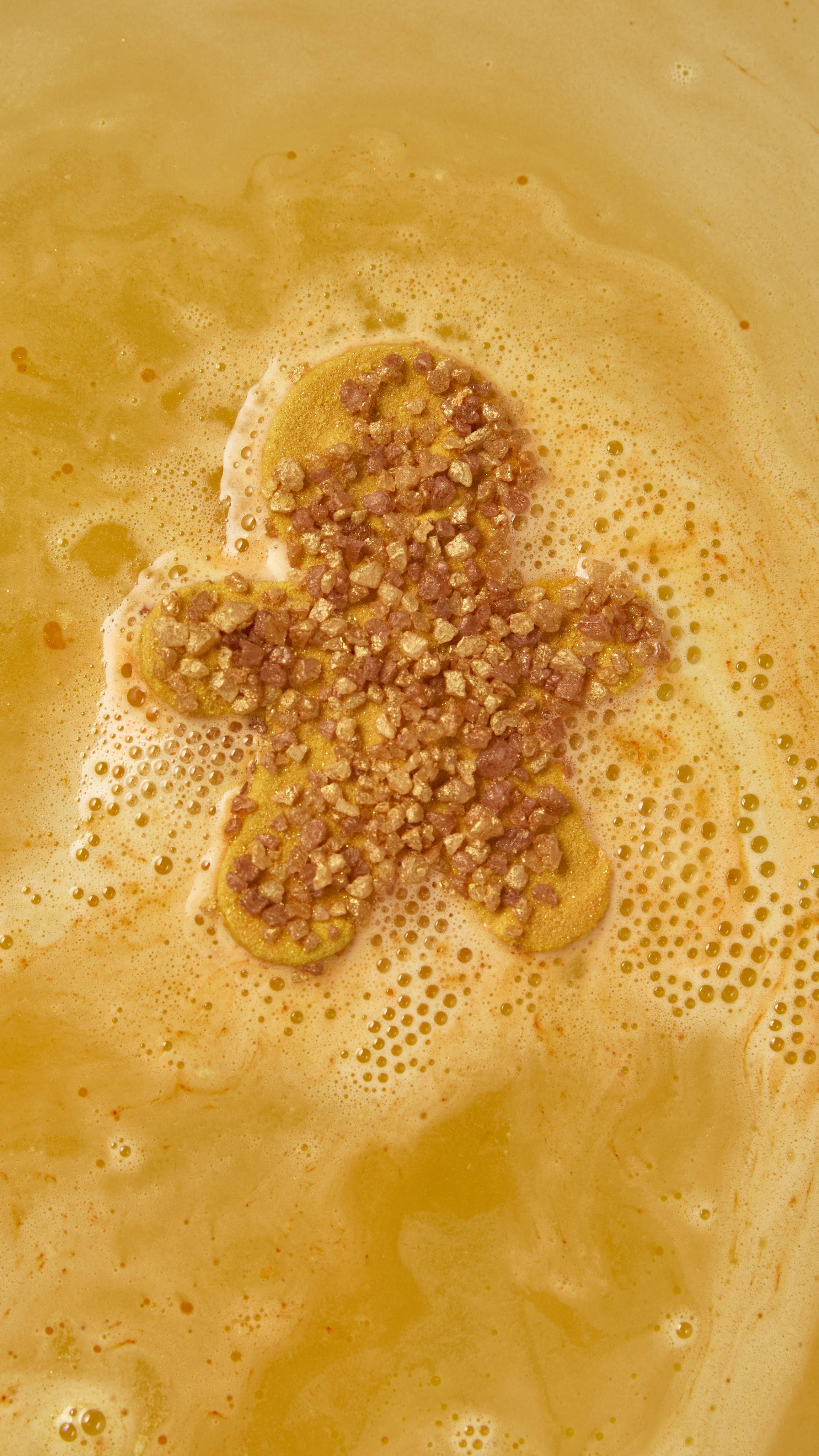 A gingerbread man-shaped bath bomb covered in golden-brown salt crystals floats on warm amber bathwater, surrounded by a border of pale golden froth and gentle bubbles.