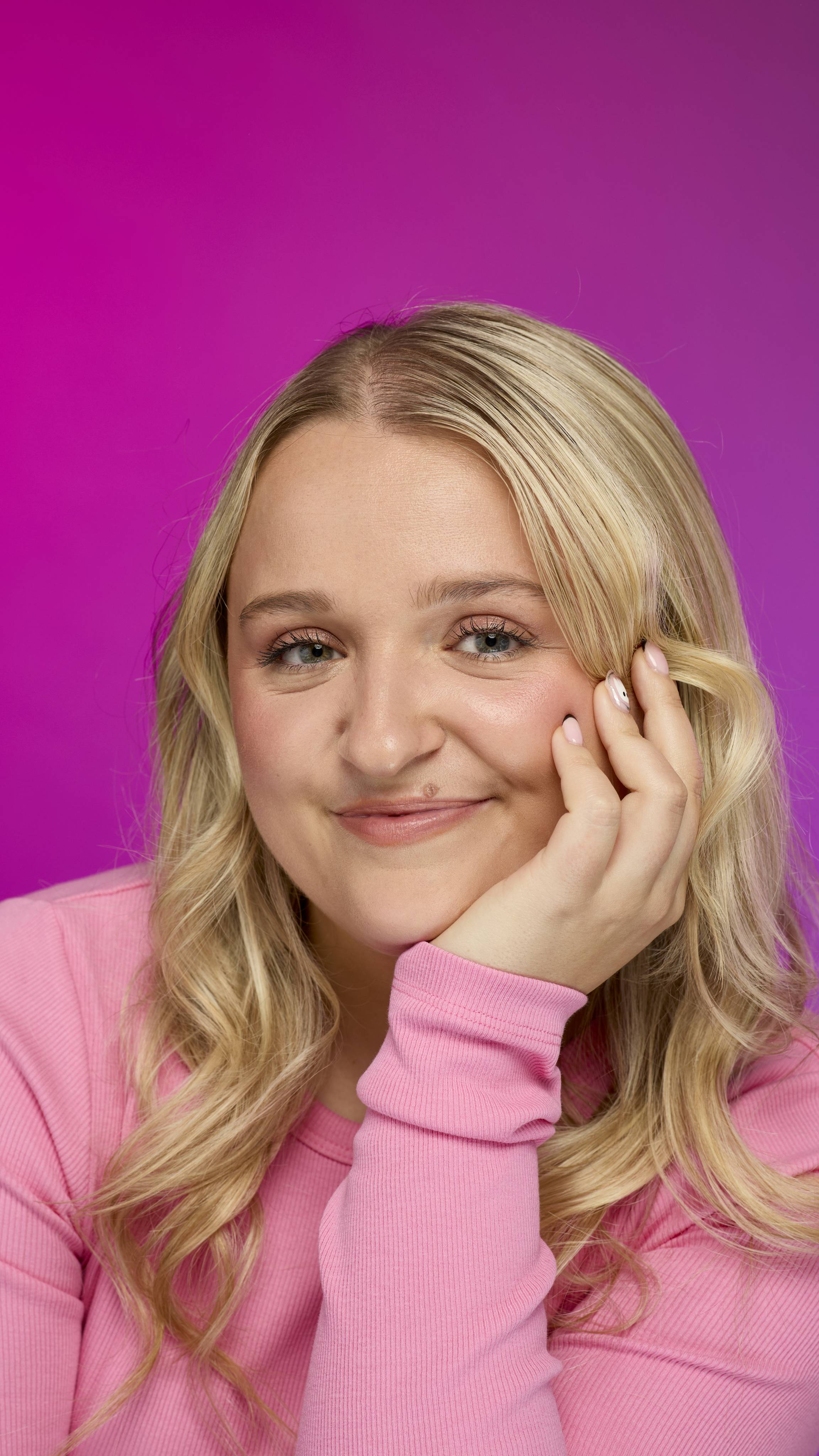 A person with long blonde hair, wearing a pink ribbed top, smiles at the camera while resting their chin on their hand. They wear a light peachy-nude lip color, likely from the Glam Nudes ~ Lip square, against a bright magenta background.