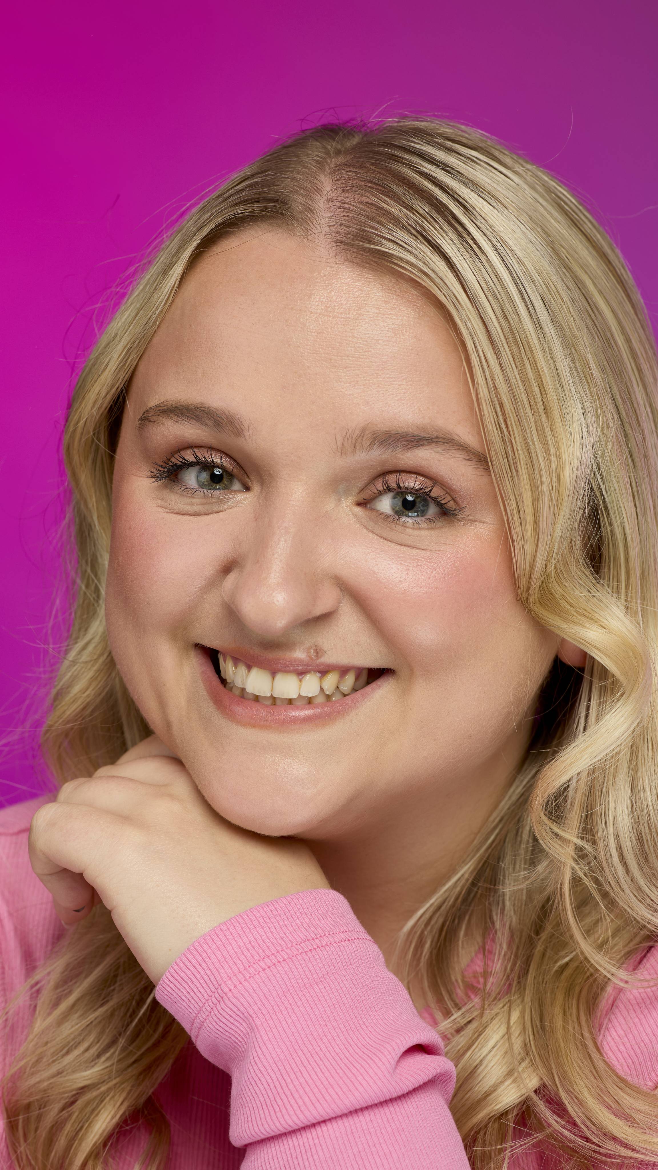 A person with long blonde hair, wearing a pink ribbed top, smiles broadly at the camera while resting their chin on their hand. They wear a light peachy-nude lip color, likely from the Glam Nudes ~ Lip square, against a bright magenta background.