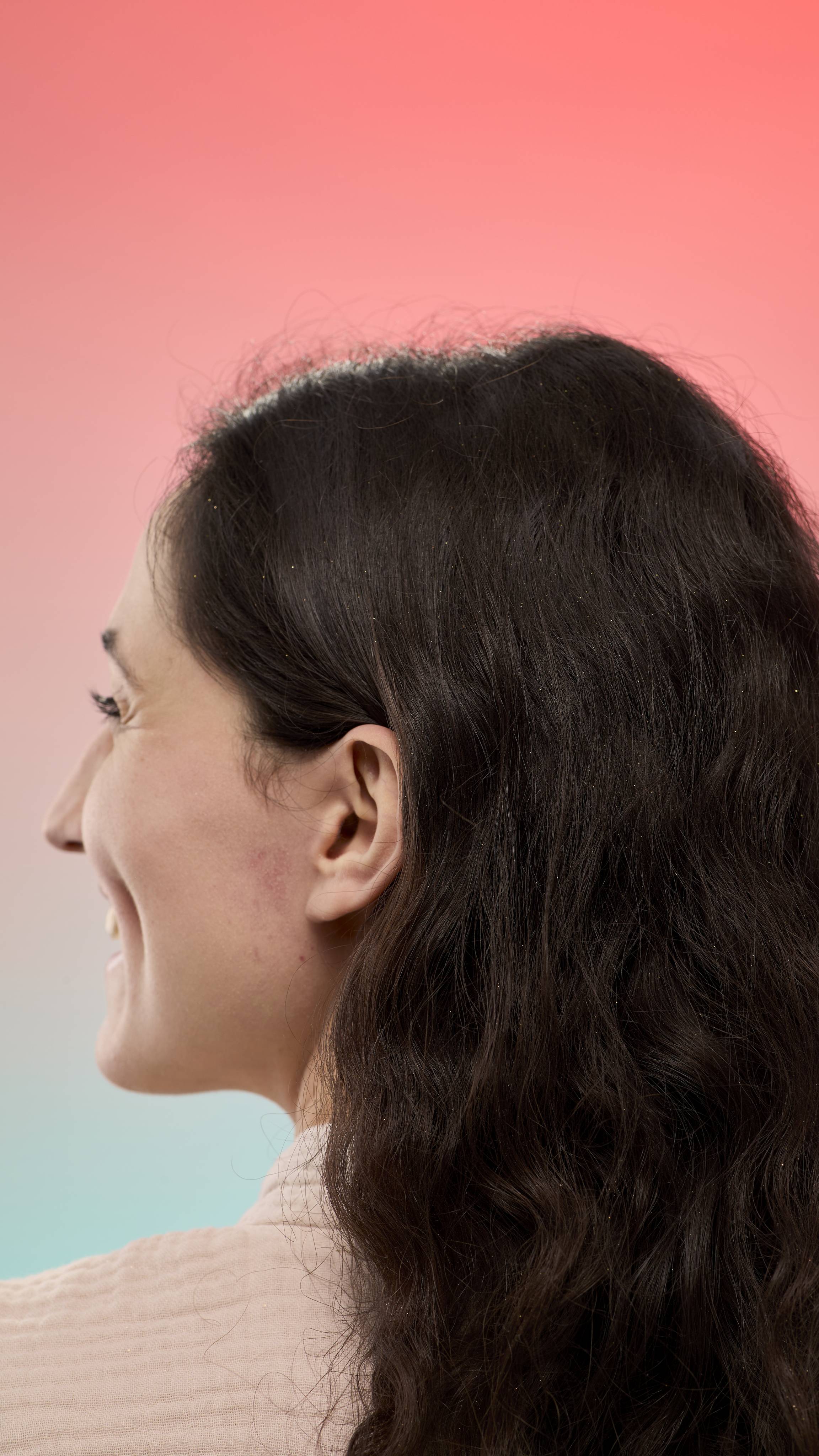 A person with dark wavy hair shimmering with fine glitter from Glam Locks dry shampoo, shown from the side. They are set against a pink-to-blue gradient background and wearing a light, textured robe.