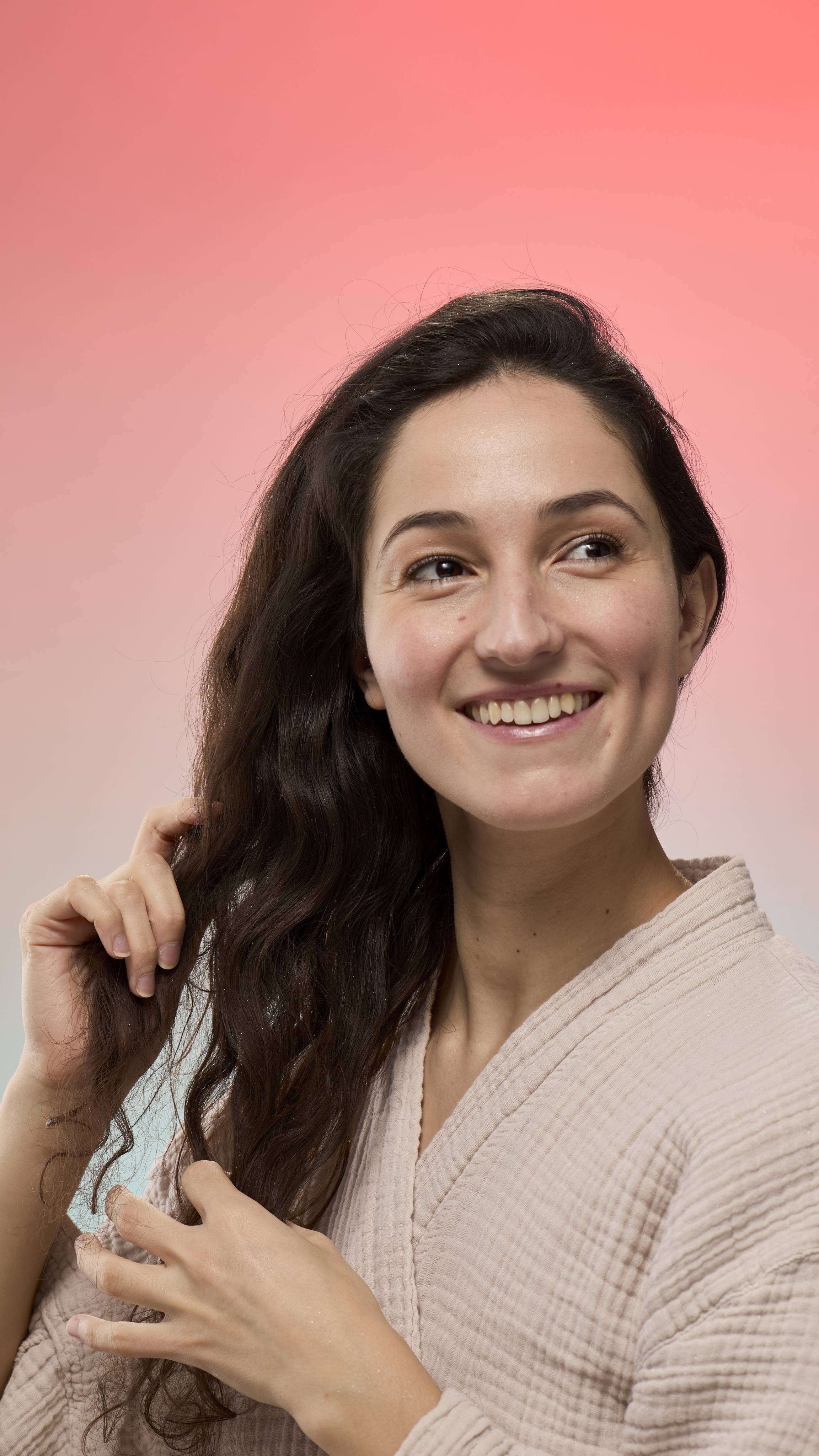 A smiling person runs their fingers through dark, wavy hair shimmering with fine glitter from Glam Locks dry shampoo. They are shown against a pink-to-blue gradient background and wearing a light, textured robe.