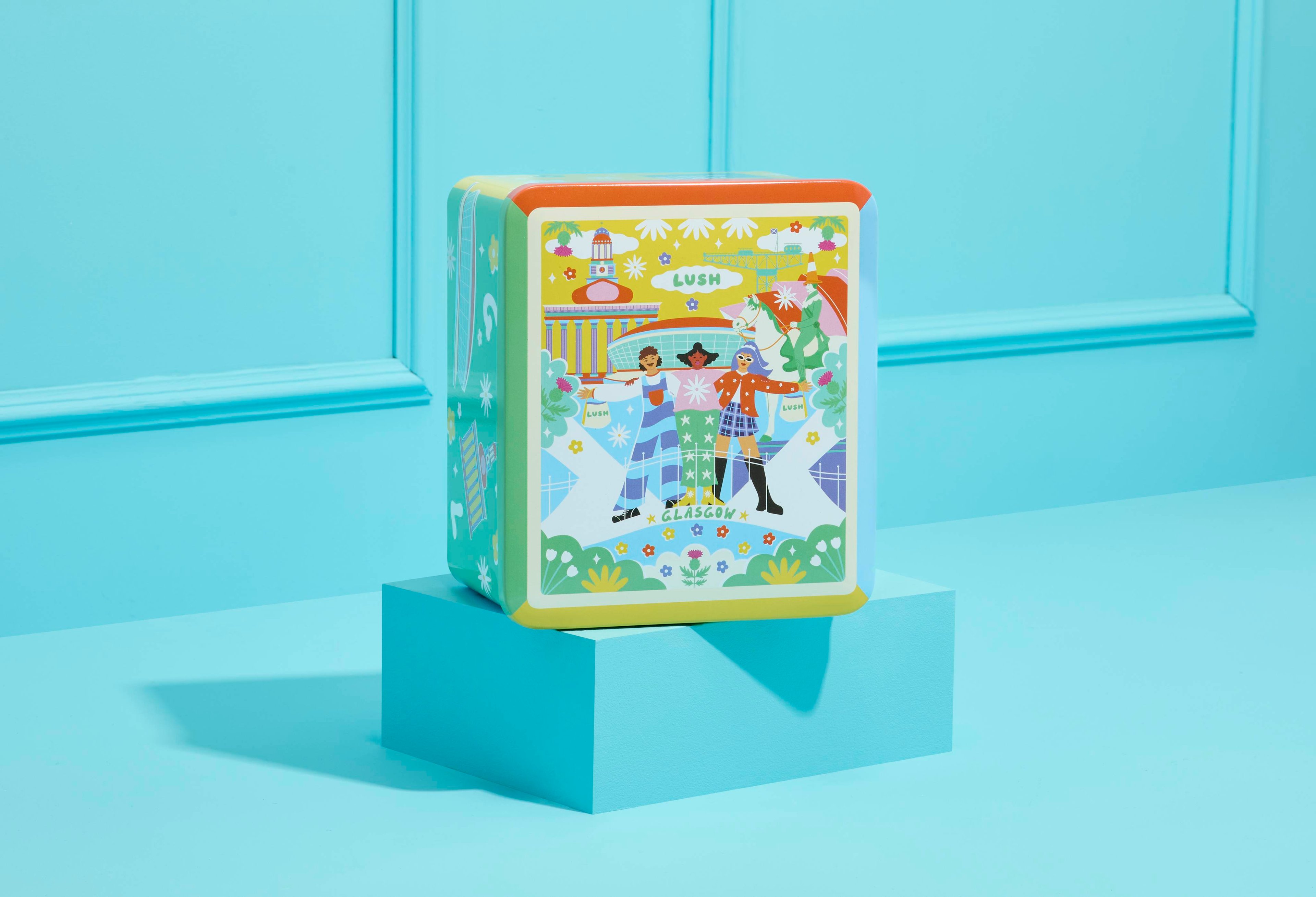 The image shows the Lush Glasgow Tin with a colourful illustration of people and landmarks on its front. The tin rests on a blue plinth against a light blue panelled wall.
