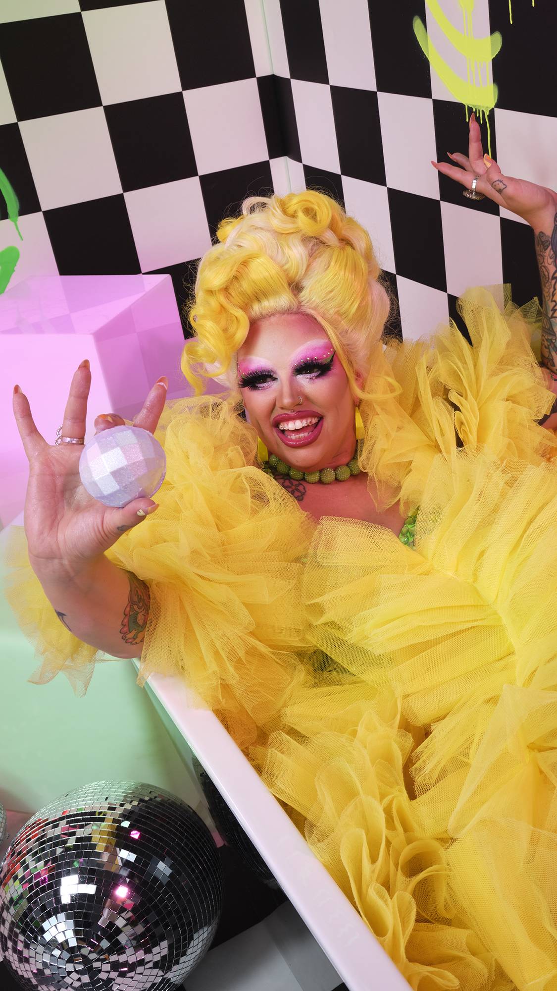 Model is wearing a yellow dress in the bathtub. They are smiling as they hold the bath bomb on checkered and neon background.