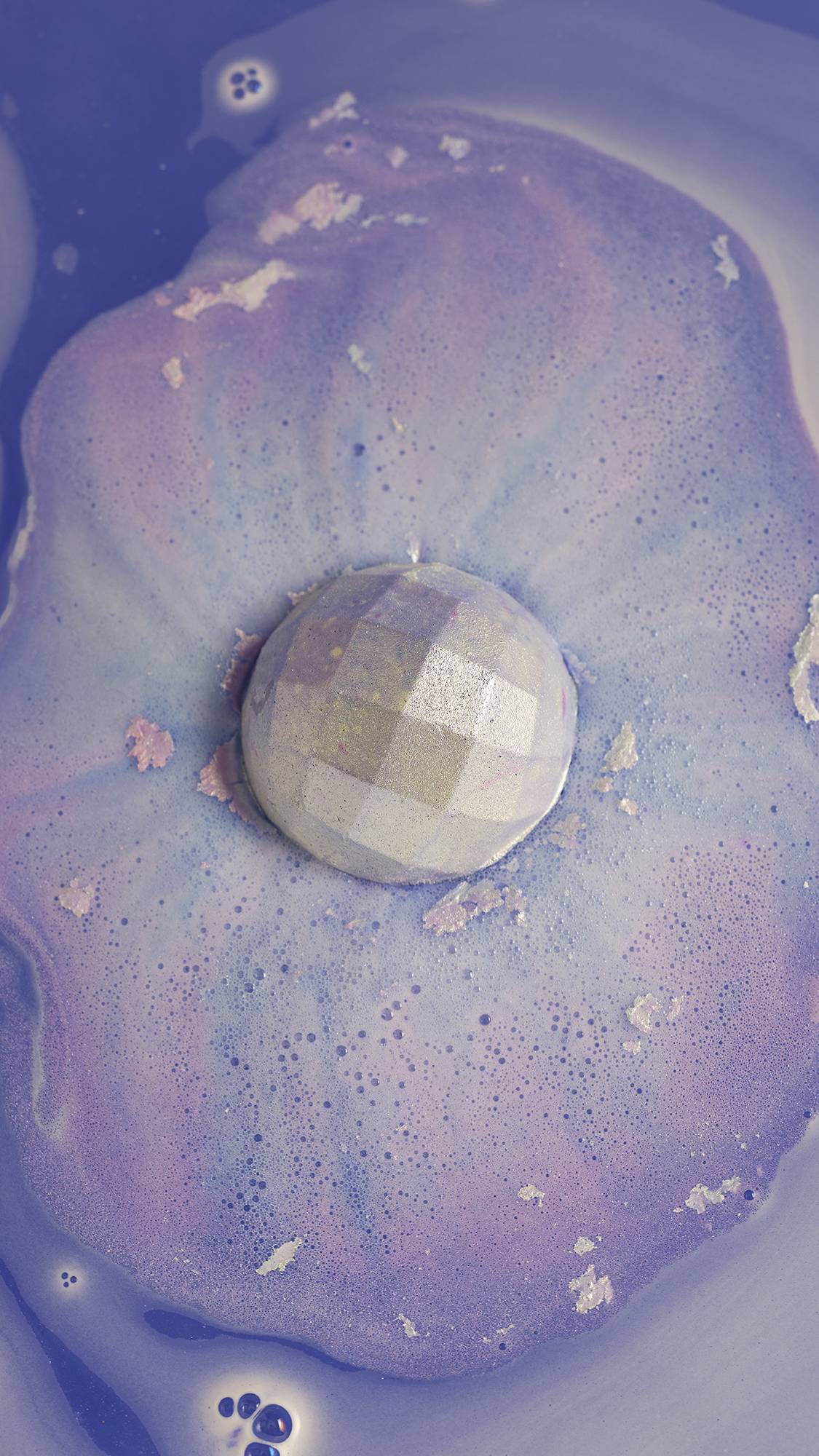 The shimmering bath bomb sits dissolving among a sea of metallic purple, blue and pink foam. 