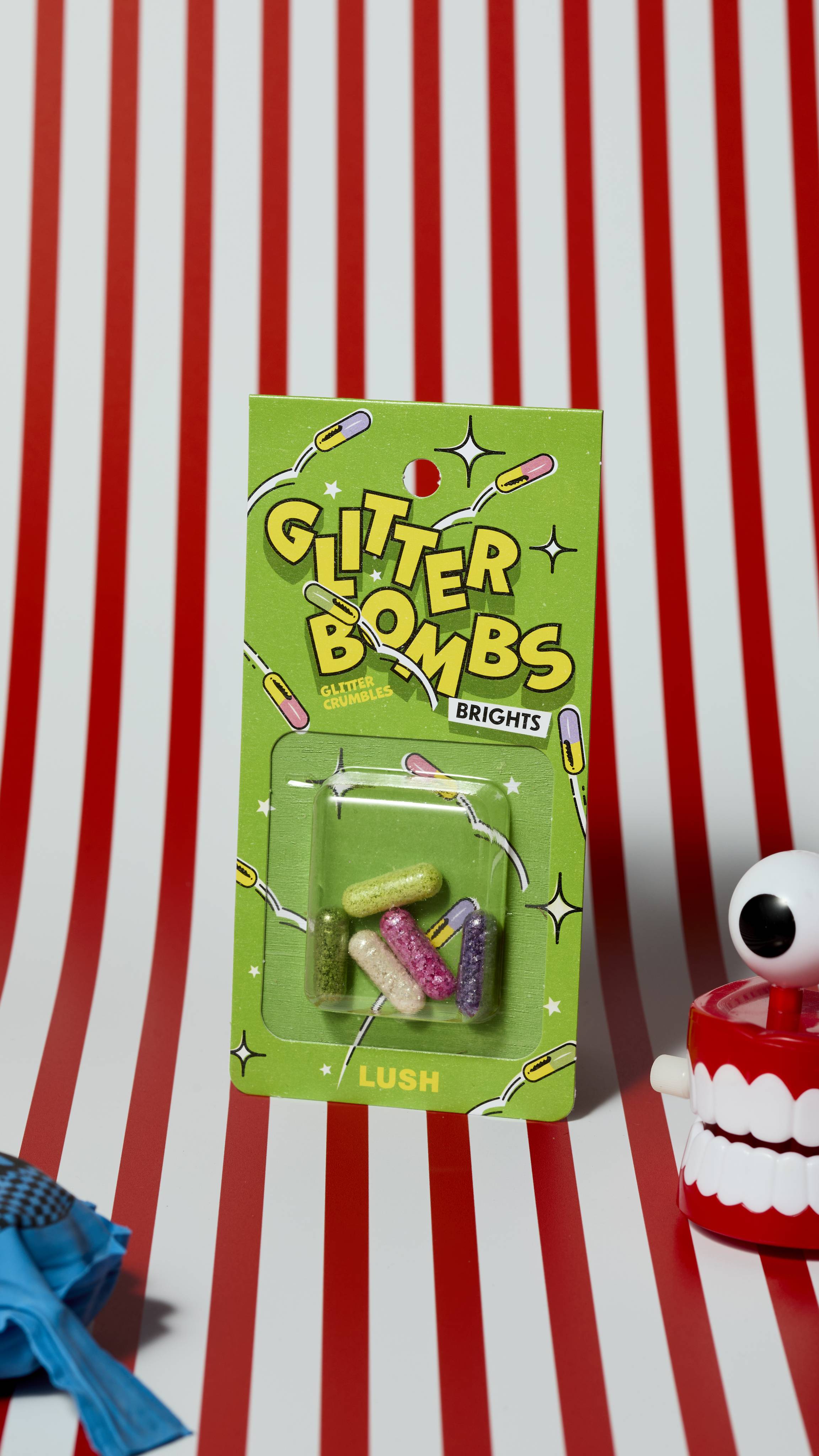 The image shows the green-packaged Glitter Bombs Bright product on a red and white, vertical striped background. There is a whoopie cushion and wind-up chattering teeth that are just visible.