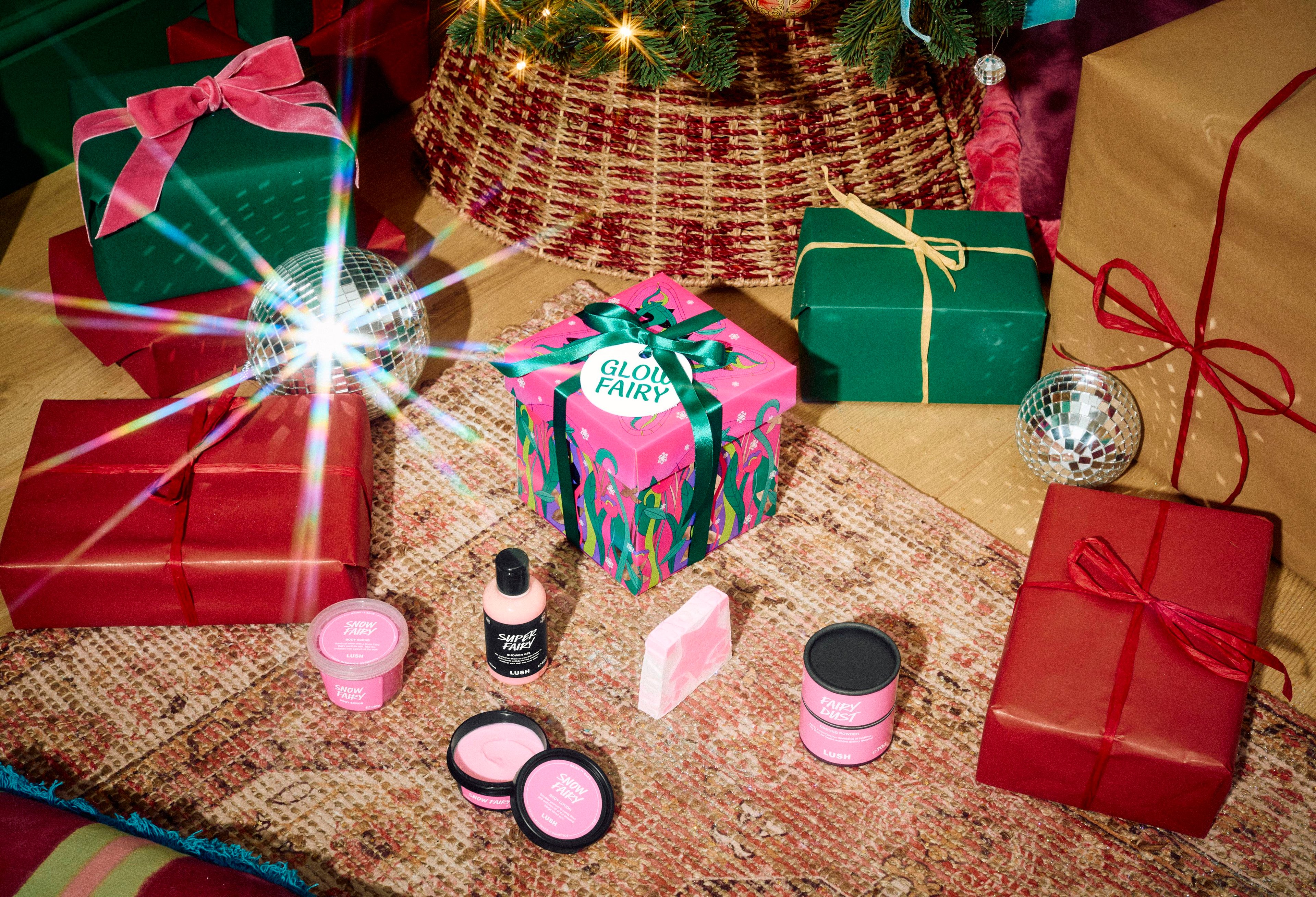 The Lush Glow Fairy gift box rests on a patterned rug with Super Fairy shower gel, Snow Fairy body scrub, lotion, dusting powder, and a soap slice, surrounded by wrapped gifts beneath a decorated Christmas tree.