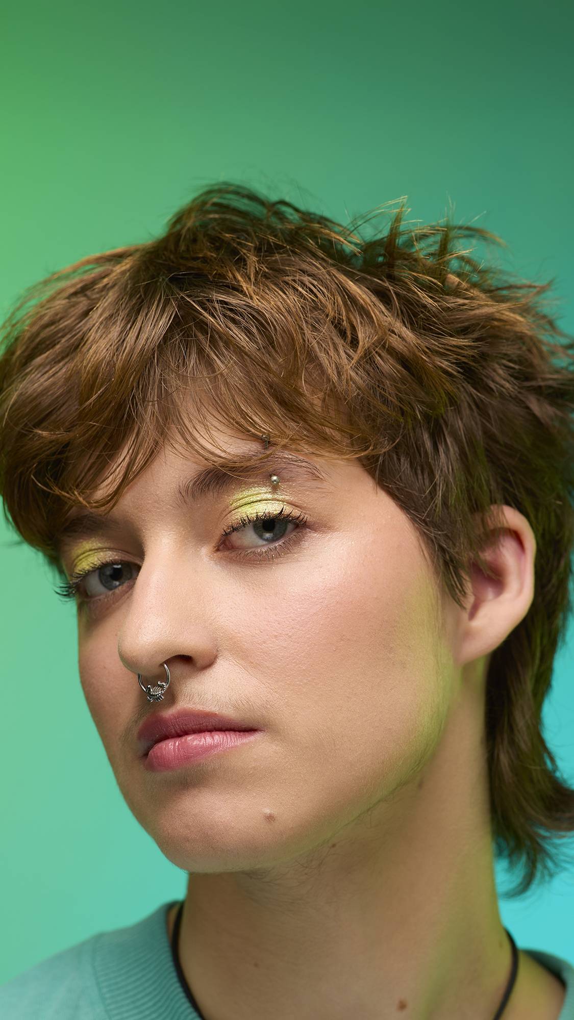 The image shows a close-up of the model's face on a pale blue-to-green gradient background. The model's make-up is done with the focal point being a delicate, lime-green, shimmering eyeshadow made by the Glow Worm Glitter Crumble. 