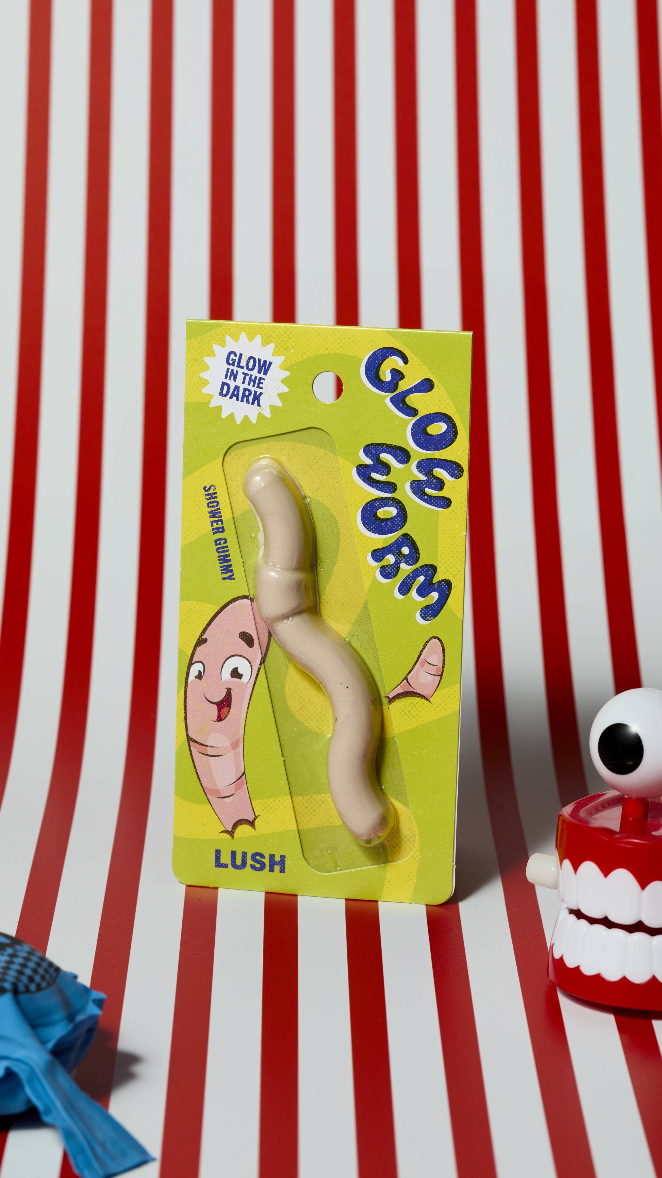 The image shows the green and yellow Glow Worm shower gummy product and packaging on a red and white striped background. There is a whoopie cushion and wind-up chattering teeth that are just visible.